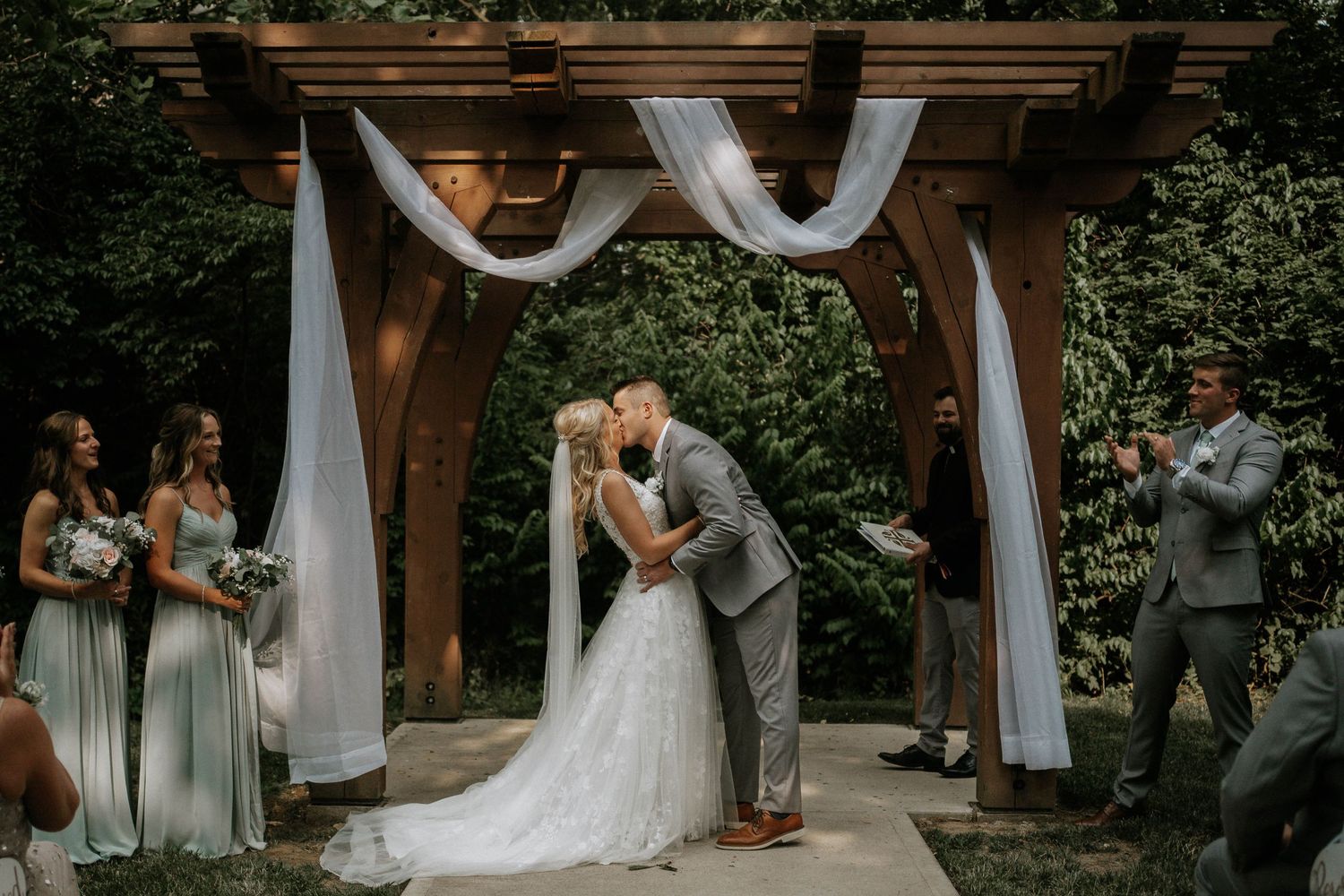 Bride and groom share first kiss under wooden pergola with white draping, surrounded by bridesmaids in sage green dresses.