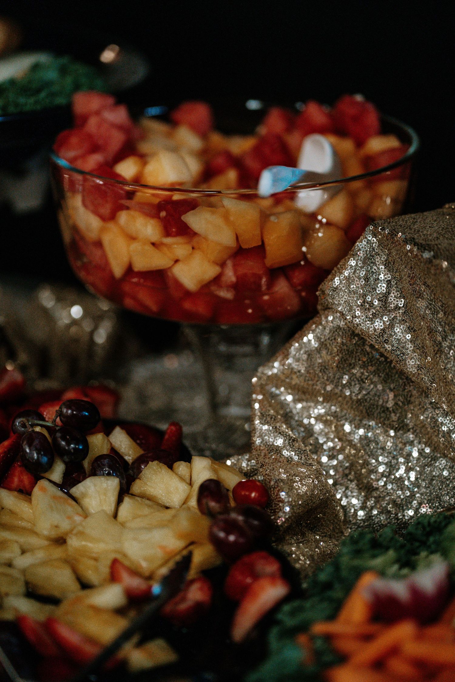 Elegant fruit salad display in glass bowl with mixed berries and melon chunks beside decorative sequined cloth.