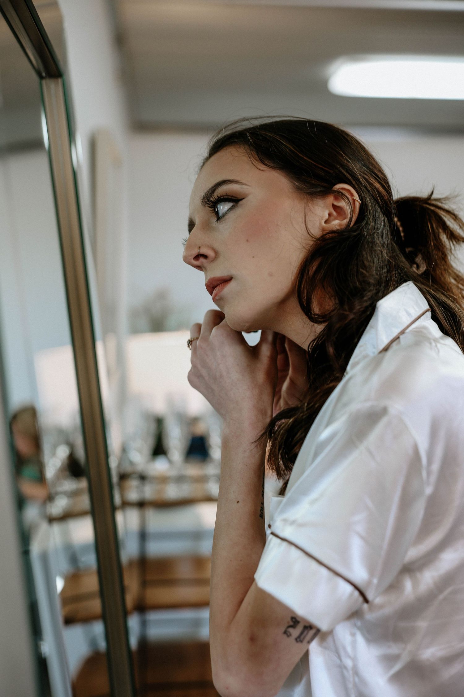 Woman in white button-up pajama shirt examining her makeup in a mirror with dramatic eye look.