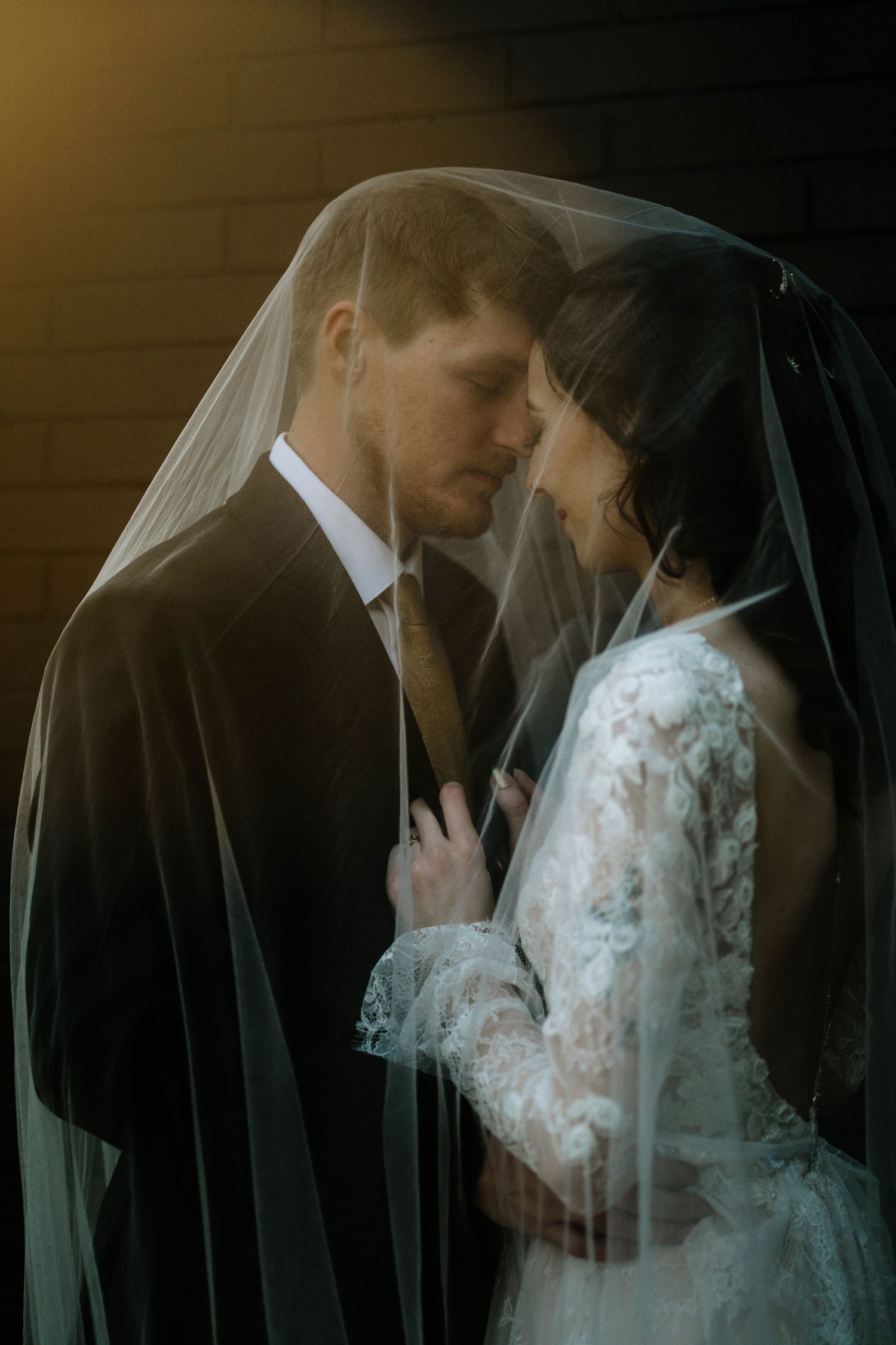 Bride and groom share romantic moment under wedding veil, bathed in warm golden light against dark brick wall.