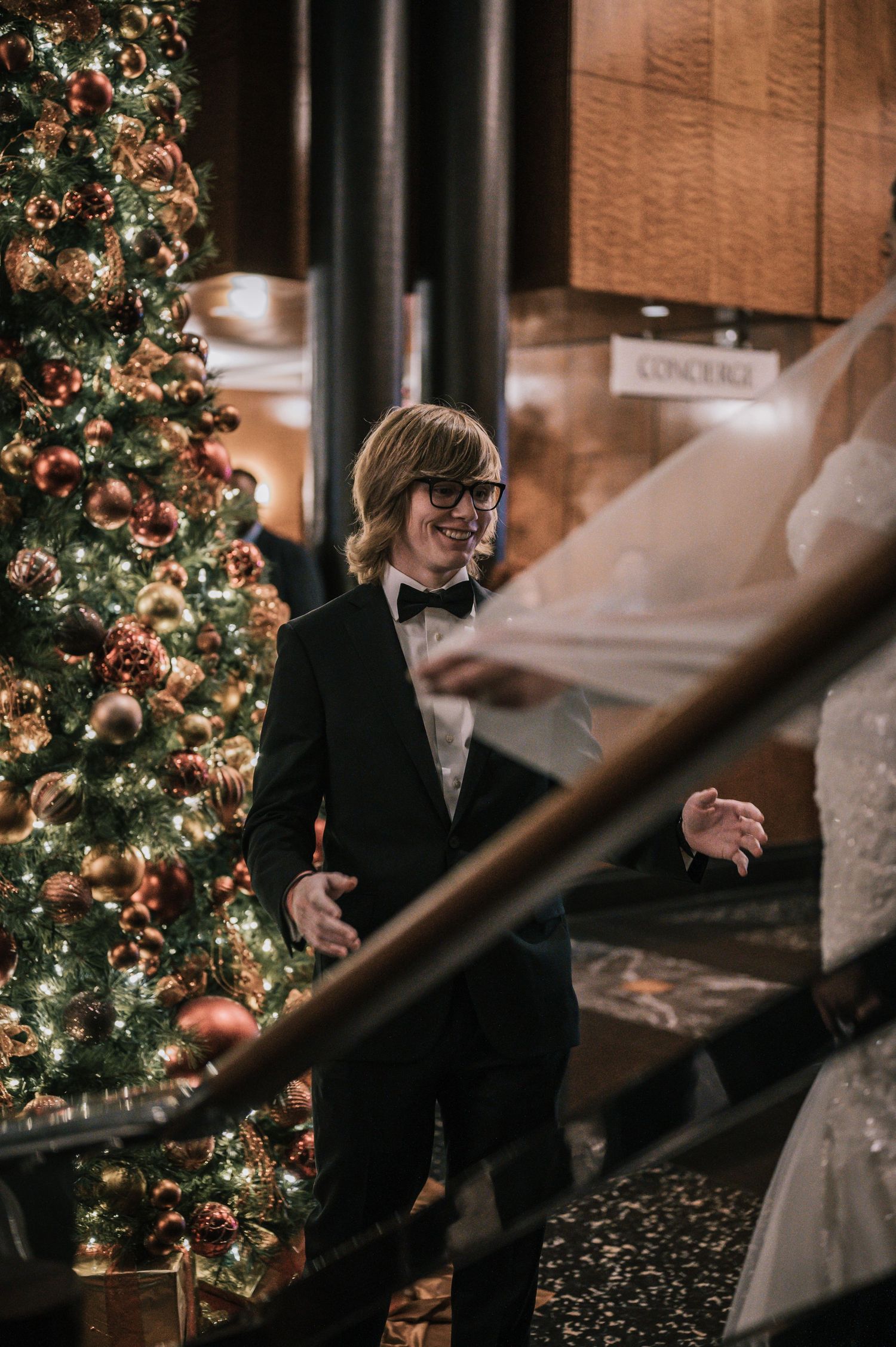 Groom in black tuxedo smiling beside decorated Christmas tree at elegant wedding venue.