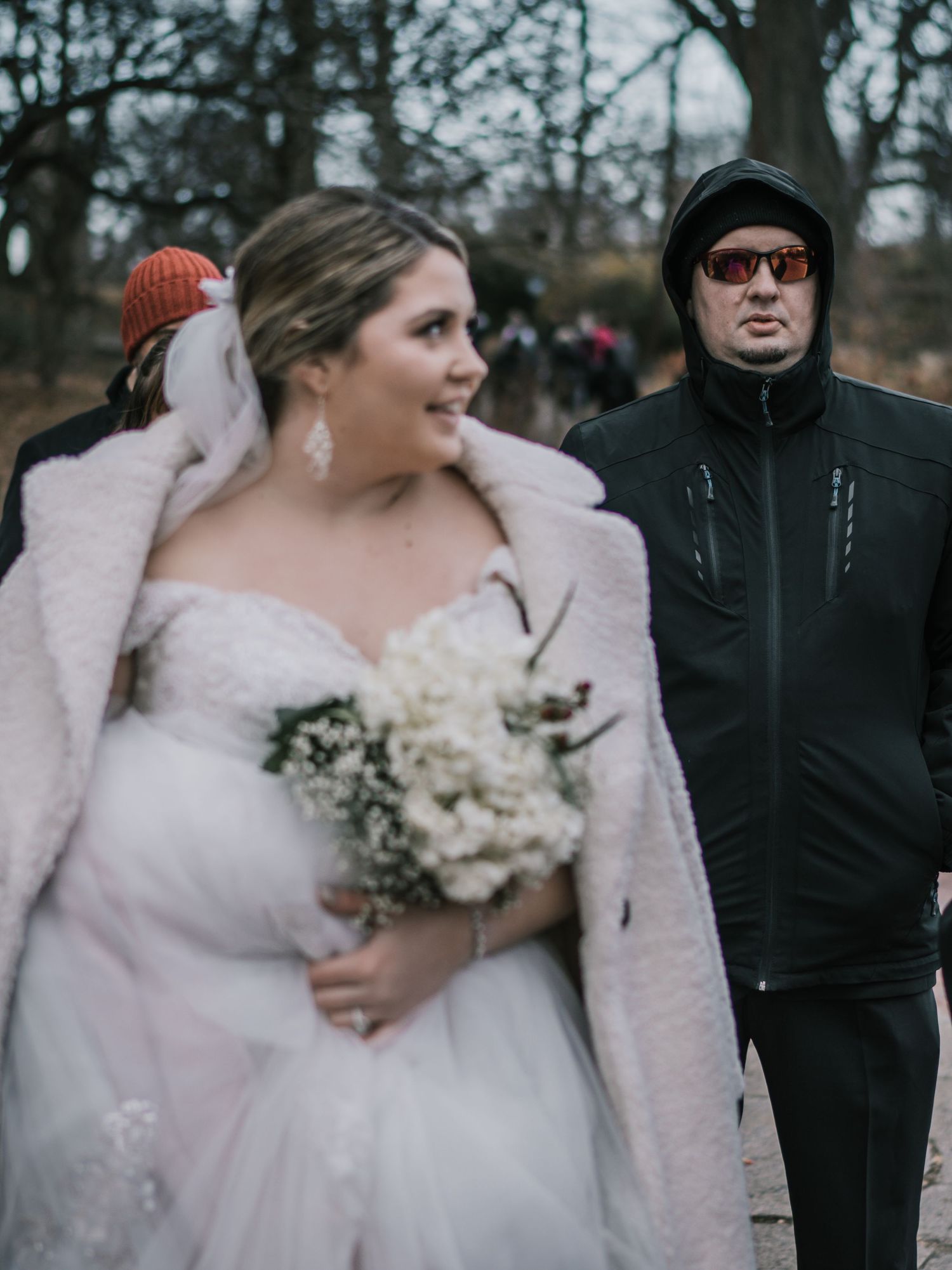 Smiling bride in white gown and fur coat holds white bouquet while walking through Central Park in winter.