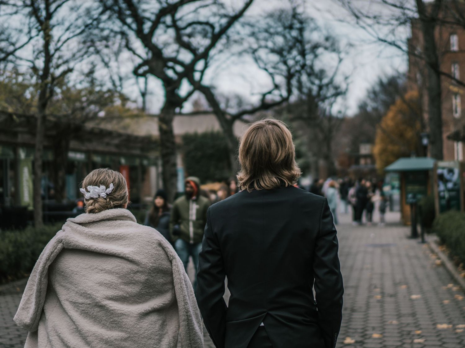 Bride and groom walk cobblestone path in autumn park, bride wrapped in cozy gray shawl.