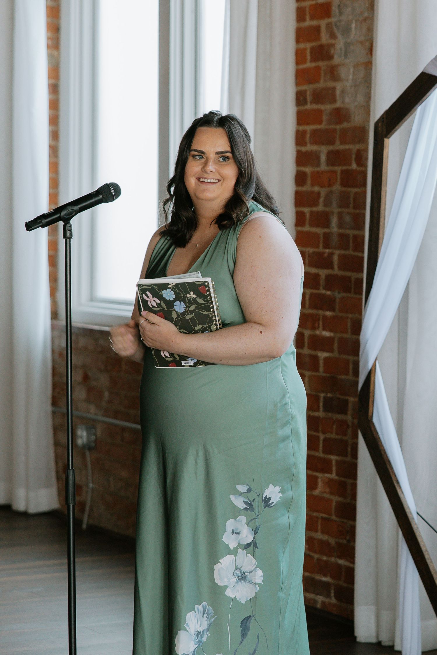 Wedding officiant in green floral dress standing at microphone holding a floral journal.