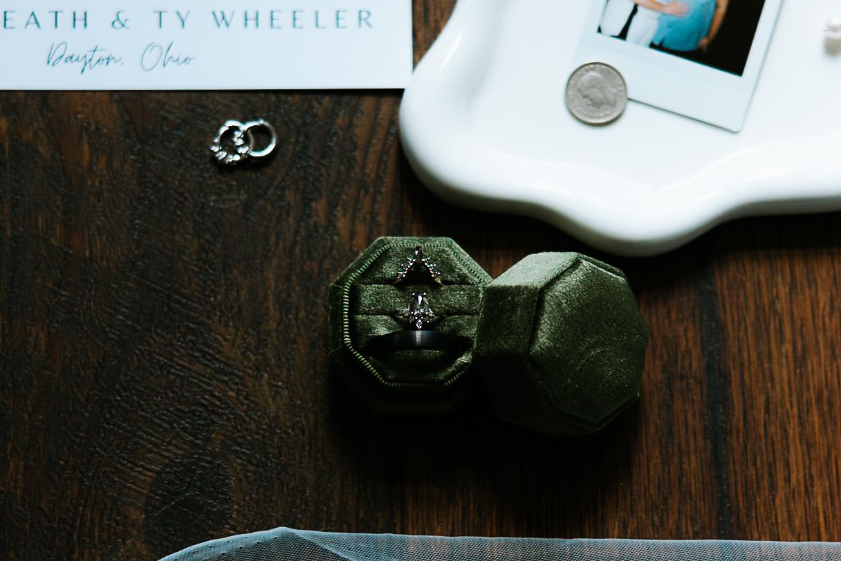 Green velvet octagonal ring box with wedding bands beside a white tray and stationery on wood.