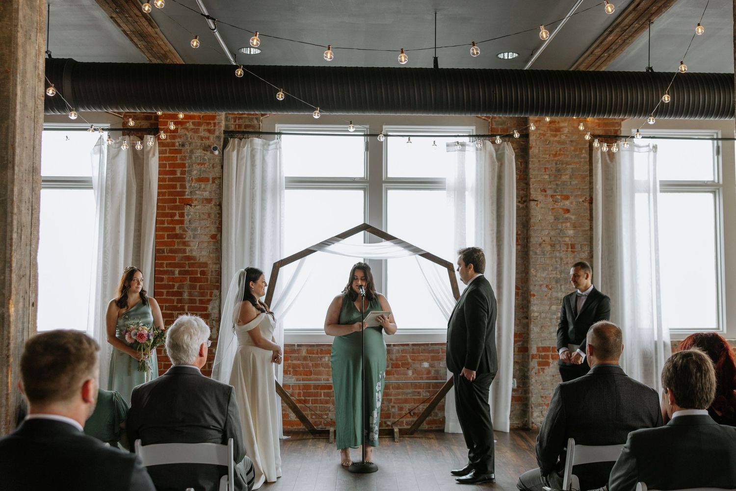 Wedding ceremony in a rustic brick venue with string lights and hexagonal arch backdrop.