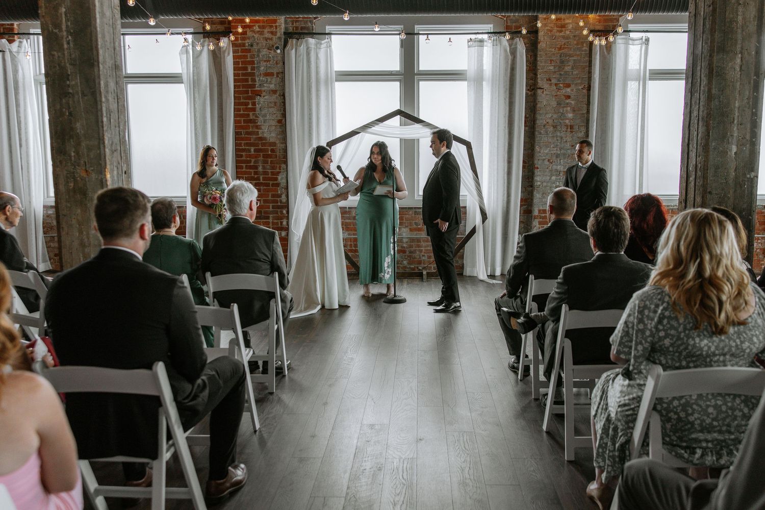 Wide shot of intimate wedding ceremony in brick venue with hexagonal wooden arch.