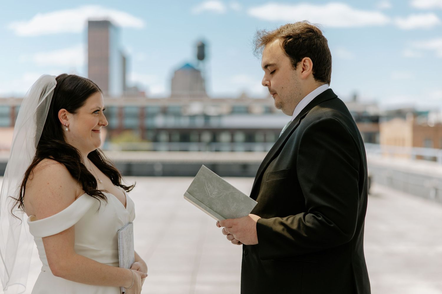 Bride smiles at groom as he reads wedding vows on a rooftop with urban skyline in background.