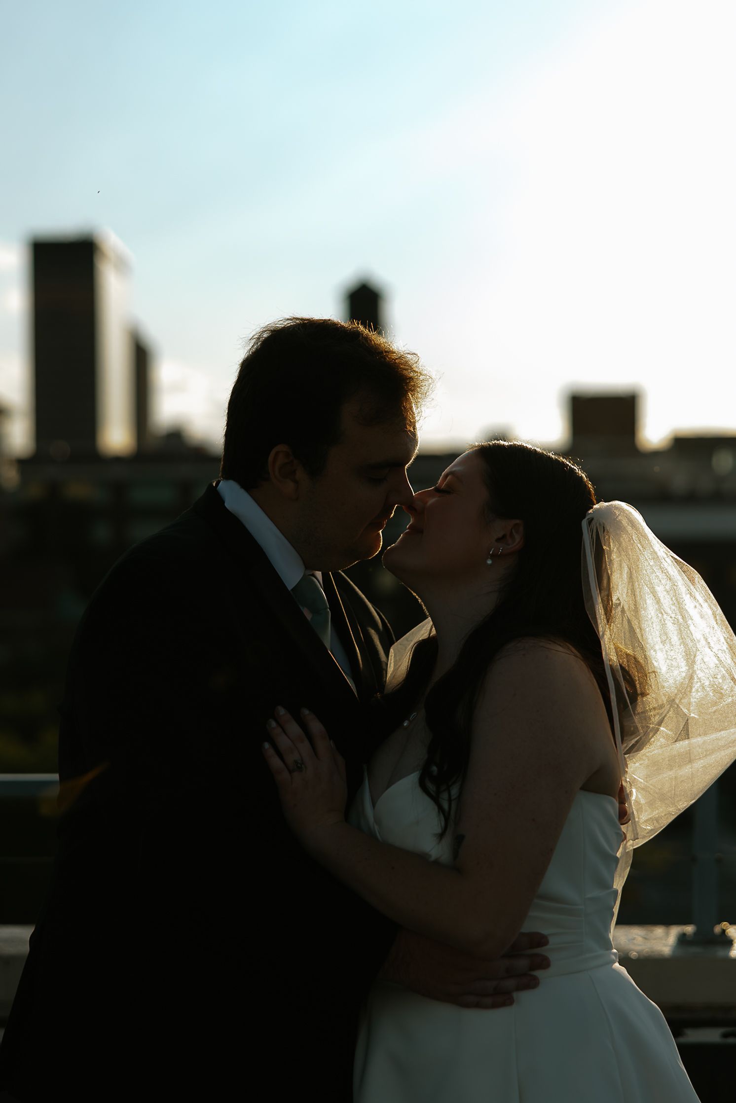Bride and groom share a romantic kiss in silhouette against a city skyline backdrop on their wedding day.