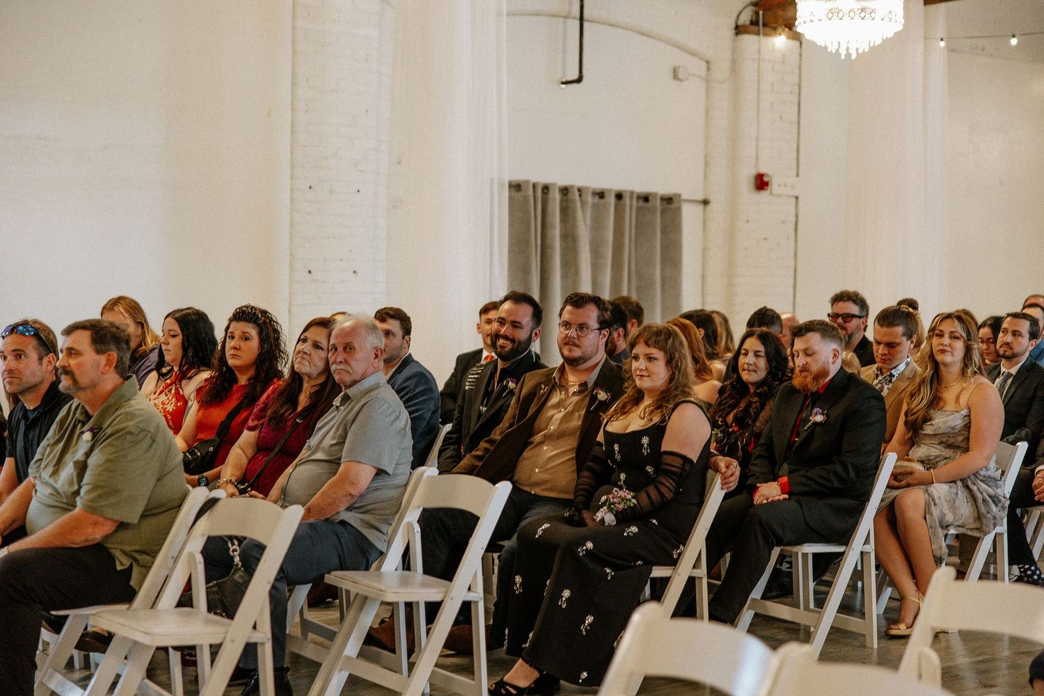 Wedding guests seated in white chairs smile and laugh during an indoor ceremony.