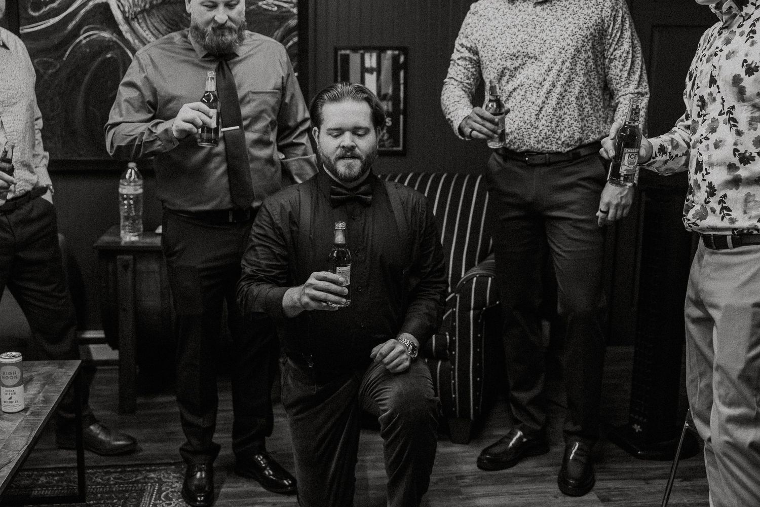 Black and white photo of a man kneeling with a drink surrounded by groomsmen.