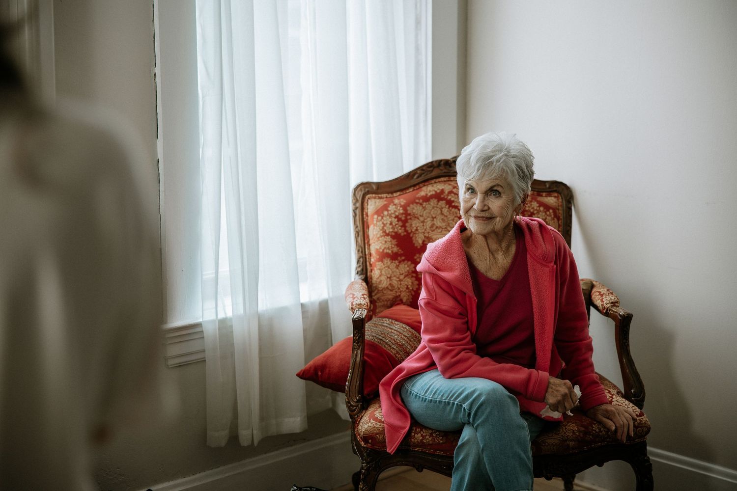 An elderly woman in a red blazer sits thoughtfully in an ornate chair near a bright window at Urban Artifact in Cincinnati, Ohio