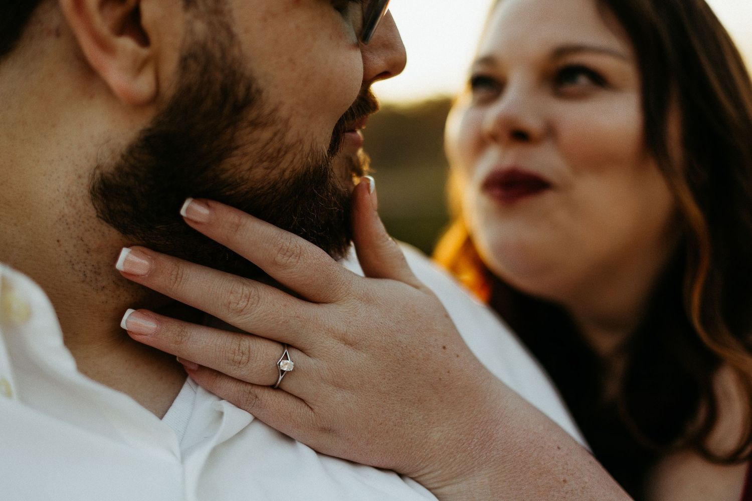 Close-up of bride showing off her diamond engagement ring while touching her smiling grooms bearded face.
