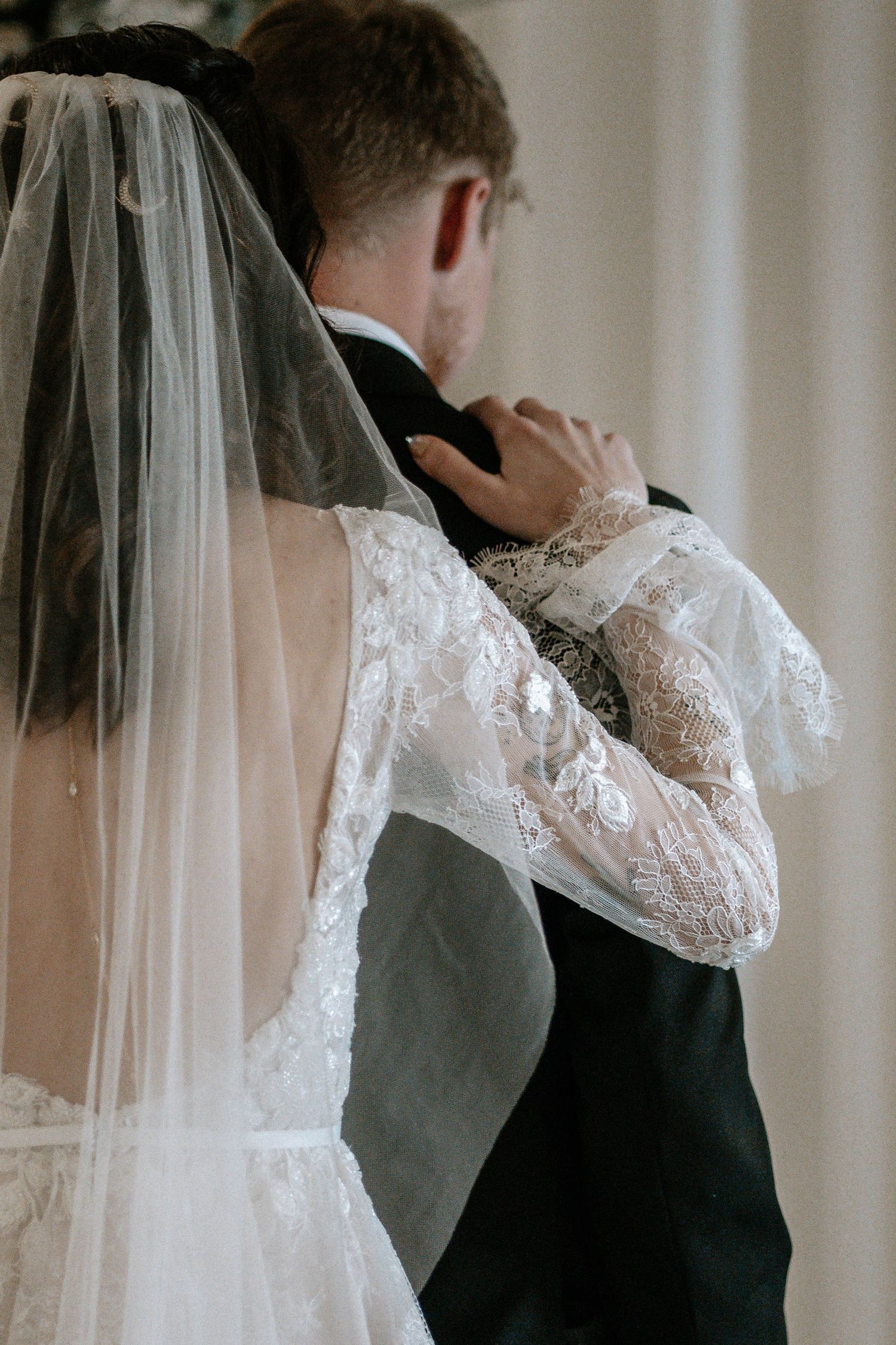 Back view of bride in lace wedding dress and veil embracing groom in black suit.