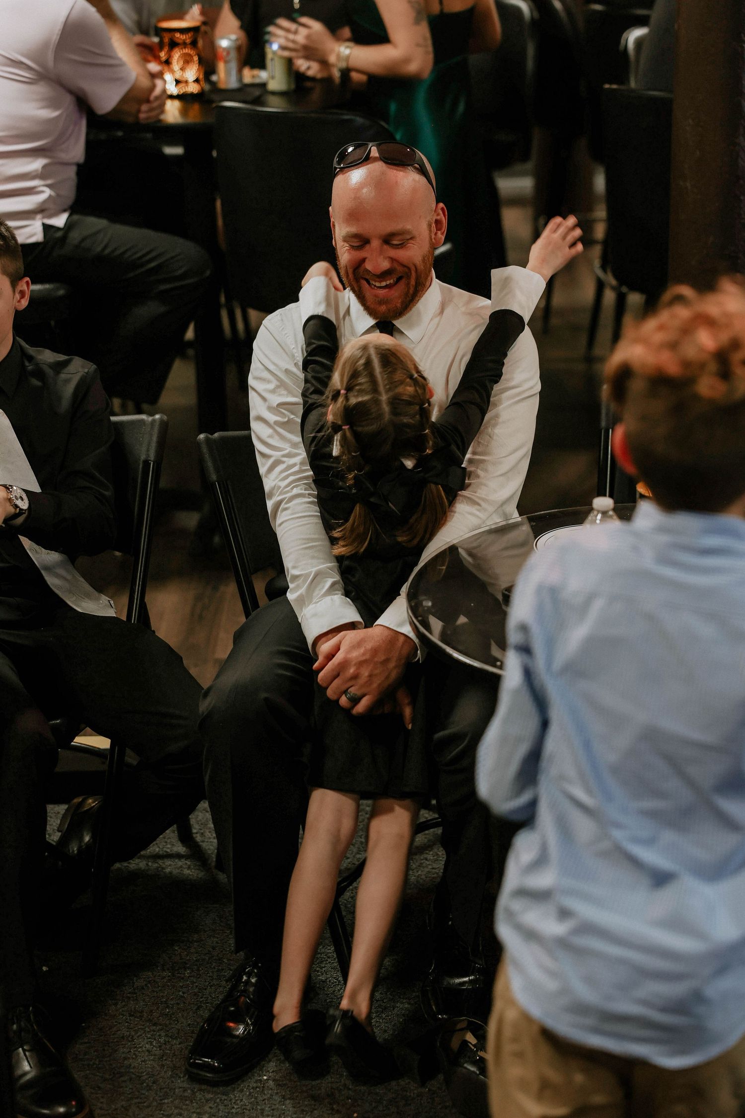A man in a white shirt laughs joyfully as a young girl hugs him at a wedding reception.