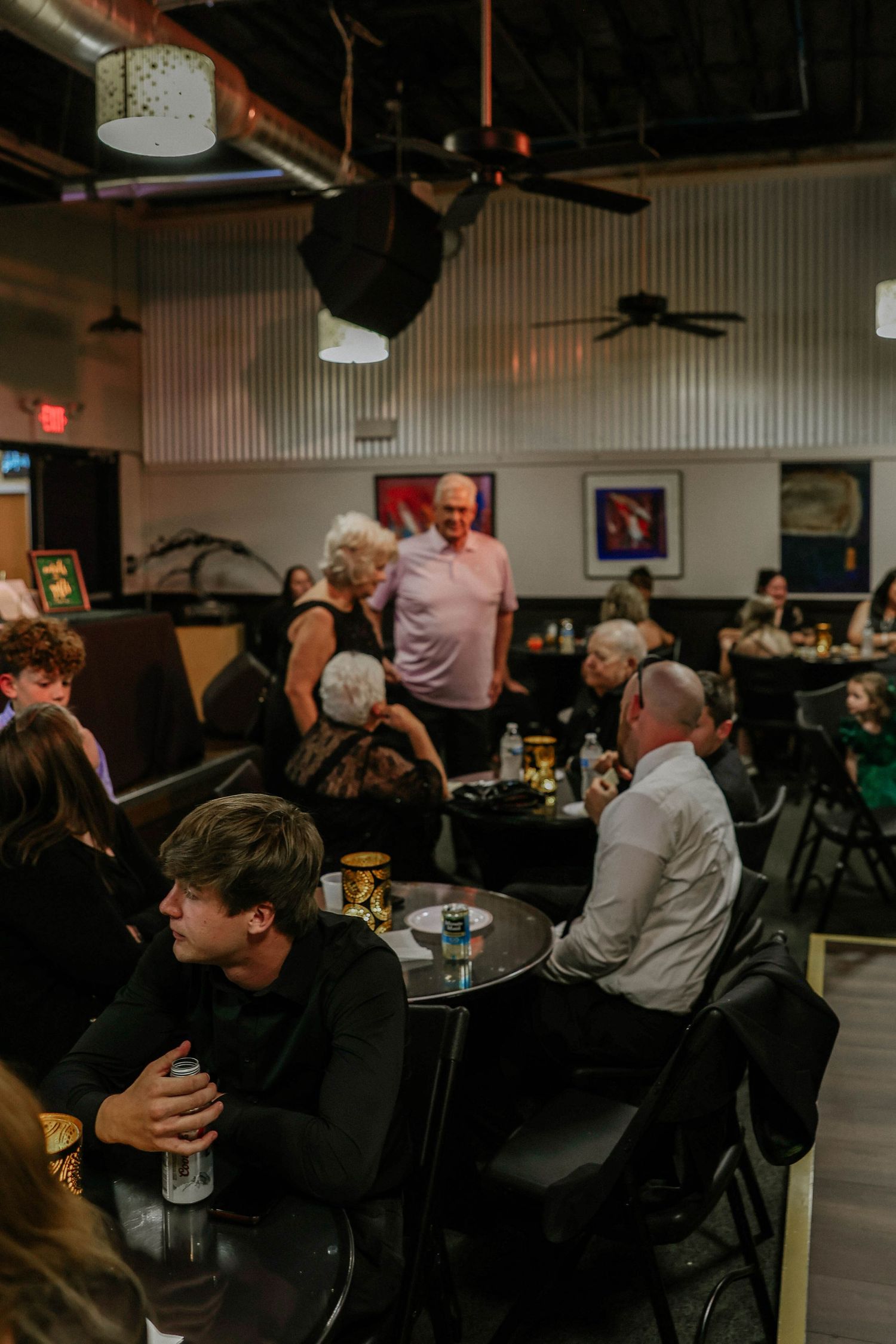 People socializing at tables in a dimly lit bar or restaurant with ceiling fans and framed artwork on walls.