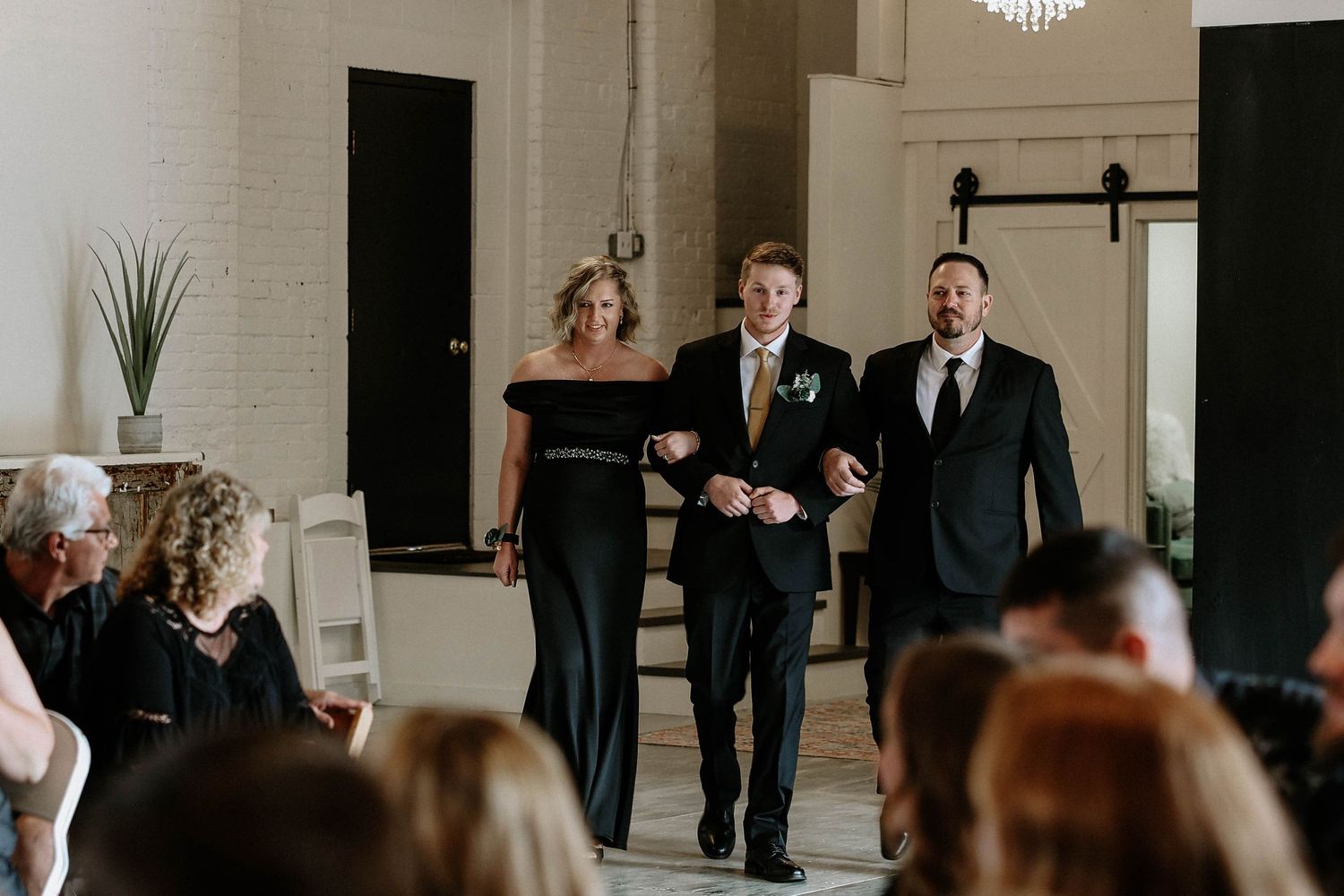 Groom escorted down wedding aisle by parents in elegant black attire at a modern white brick venue.