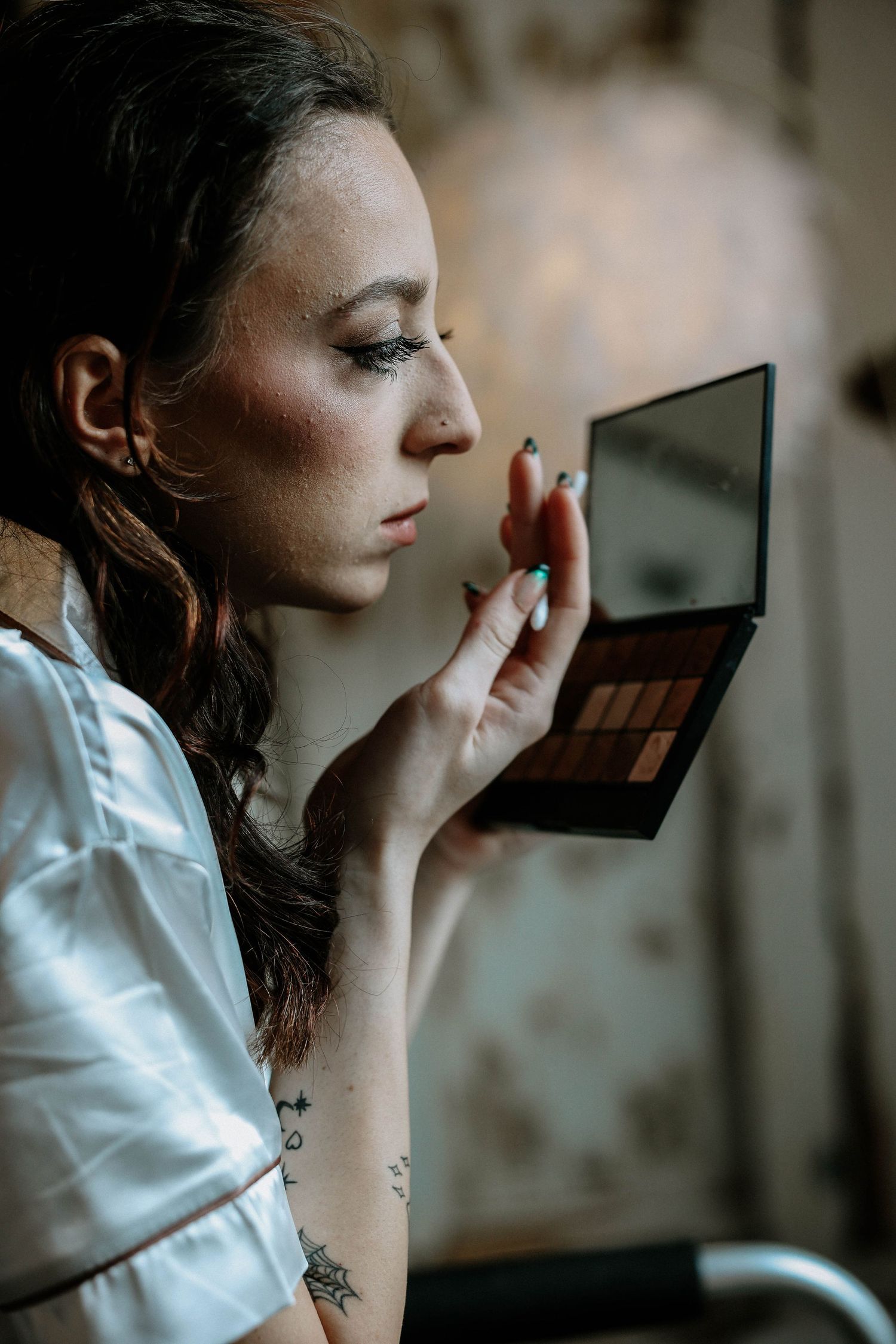 A bride applies eyeshadow using a compact mirror and brush during wedding day makeup.