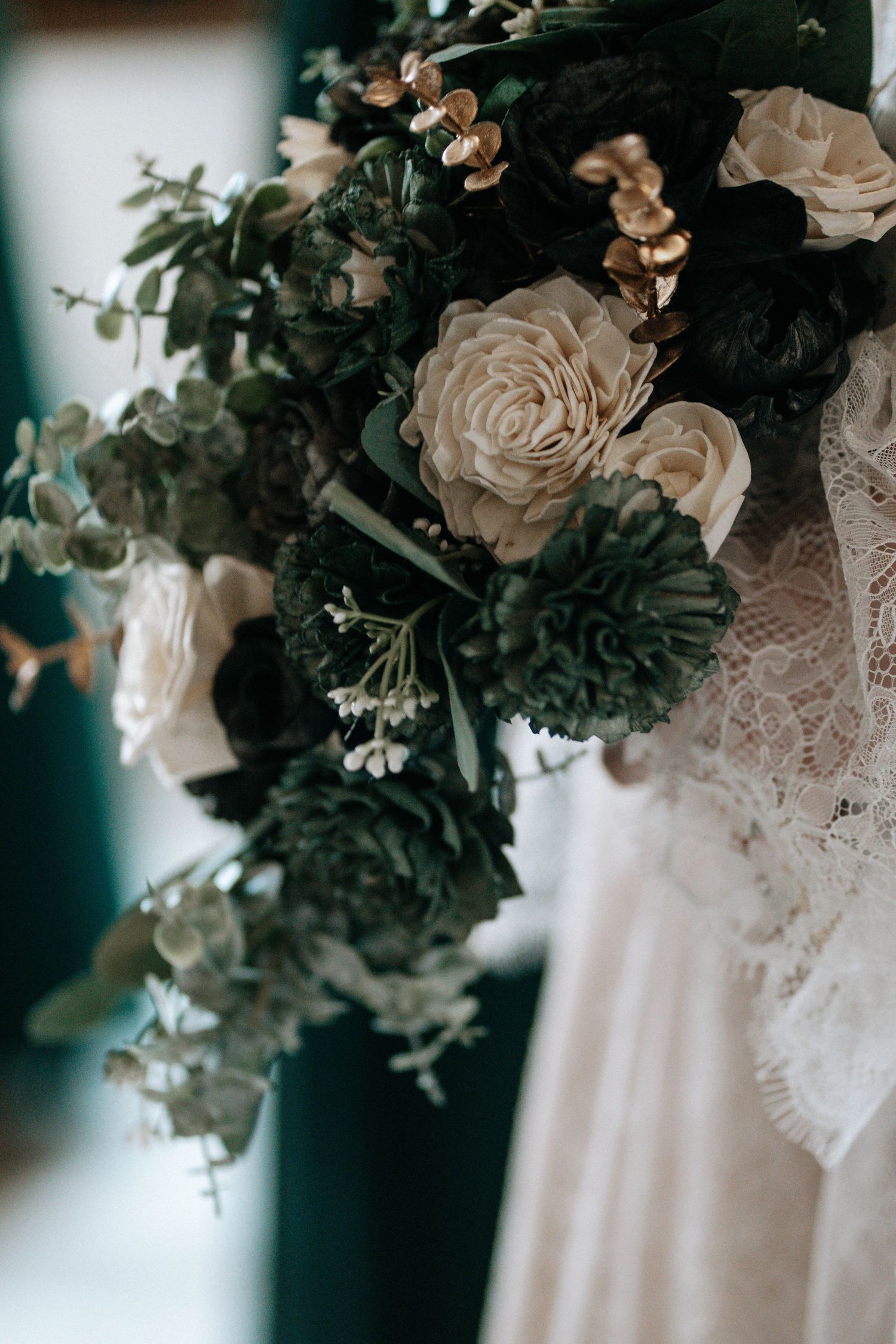 Close-up of elegant wedding bouquet with ivory roses, dark green flowers, and eucalyptus next to lace bridal gown.