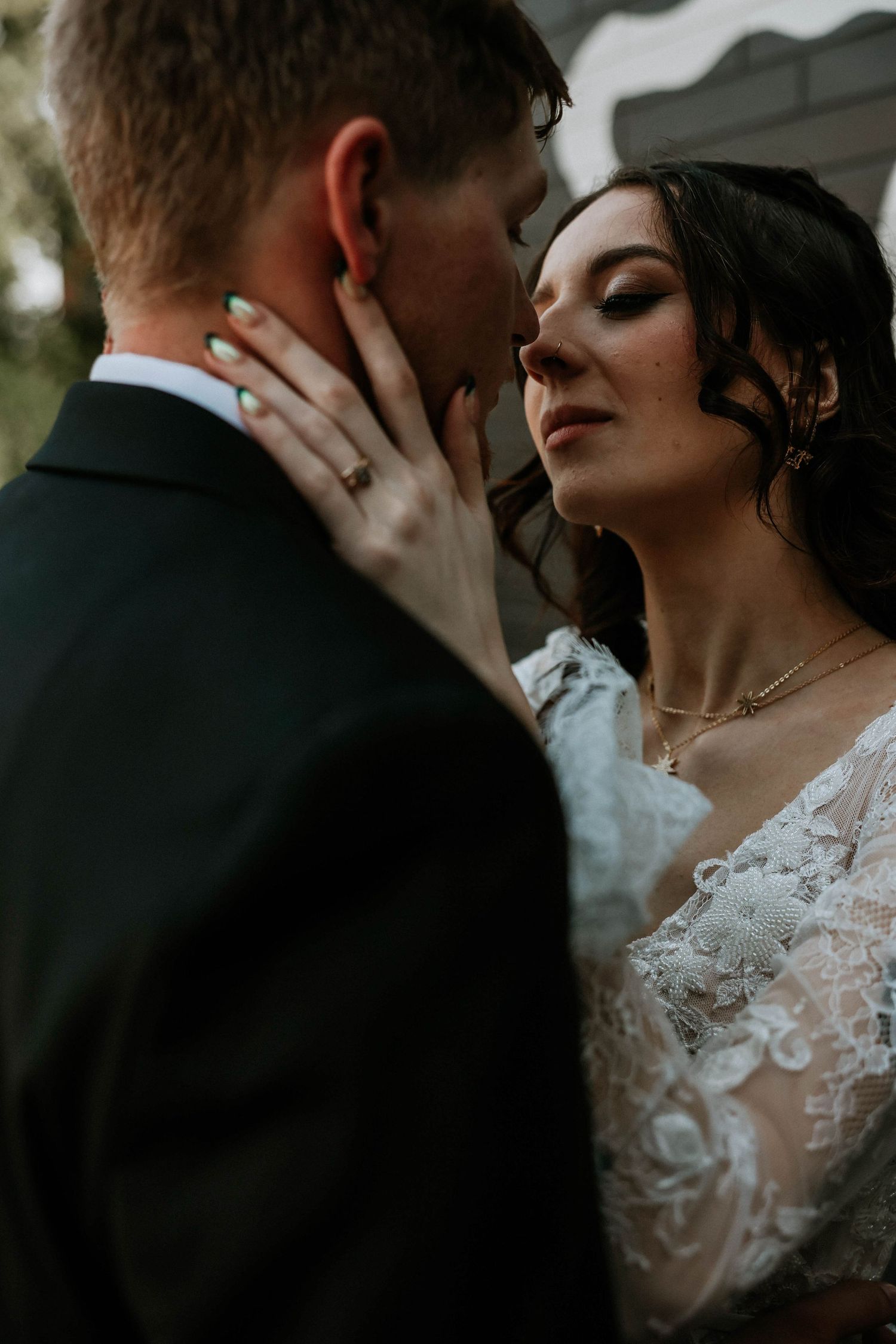 Bride in lace wedding gown tenderly holds grooms face as they share an intimate moment before their wedding kiss.