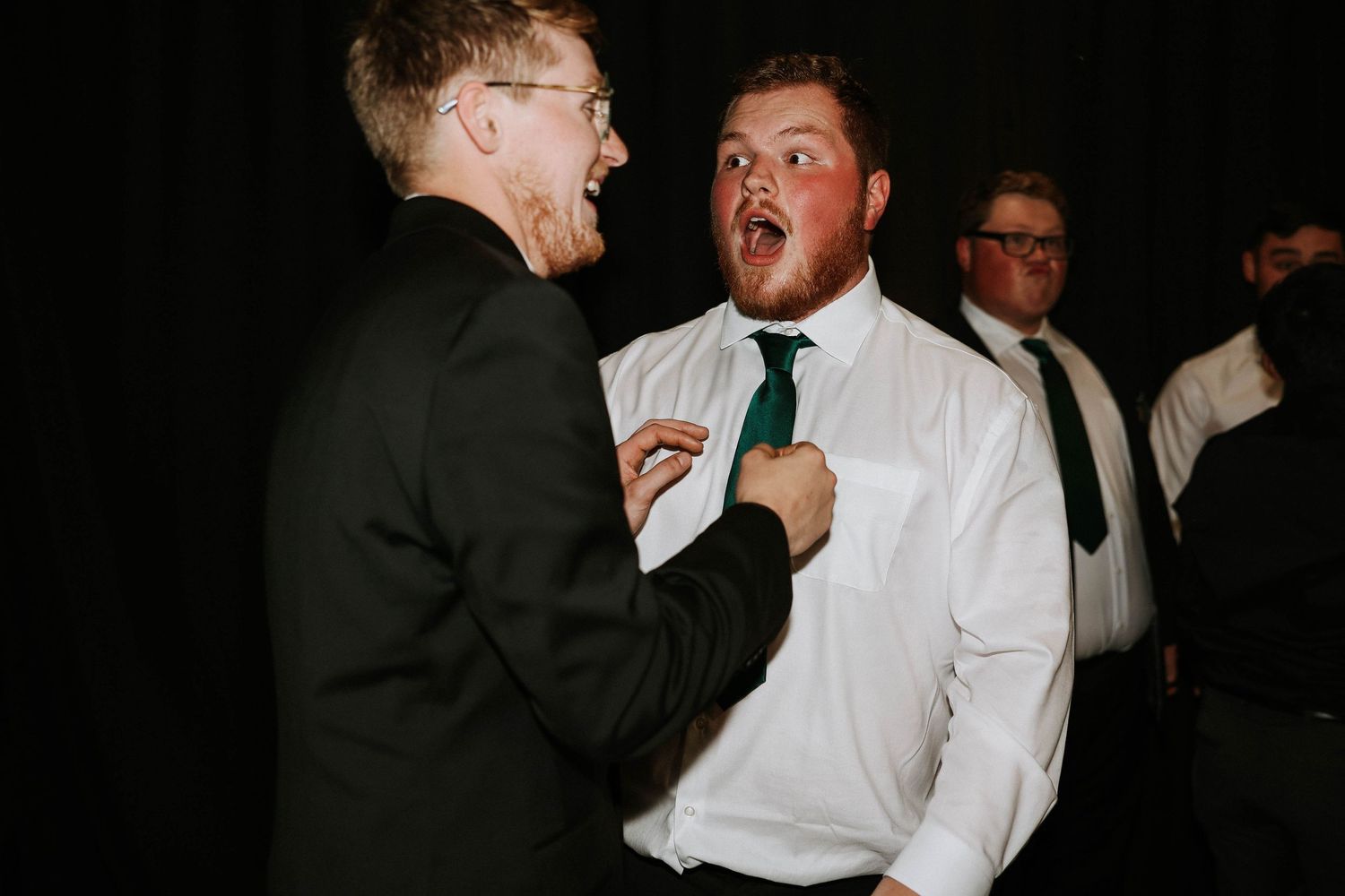 Two men in formal attire laugh and joke together at a wedding reception, one wearing a green tie.