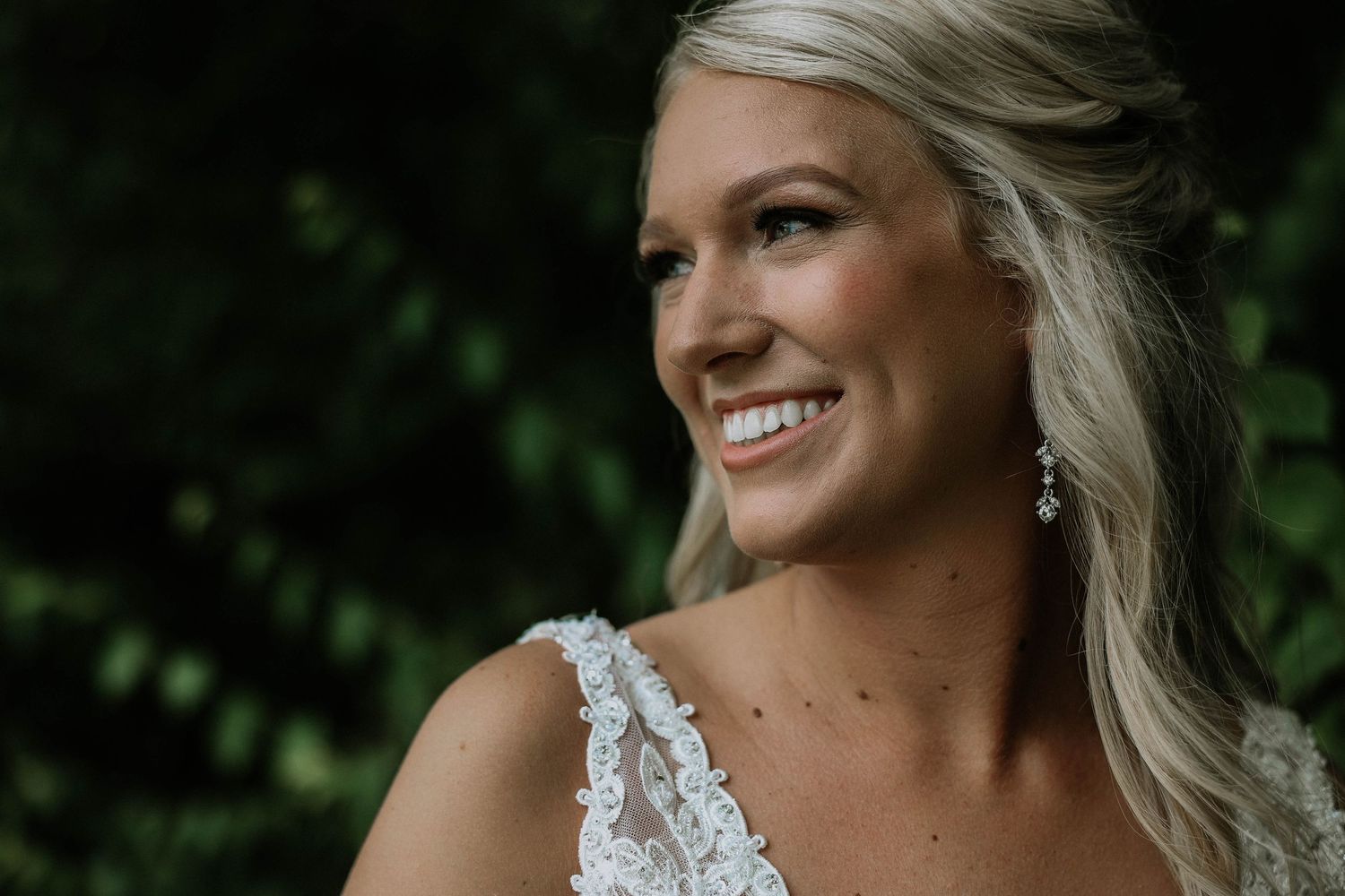 Close-up portrait of a smiling blonde bride in lace gown with drop earrings against green foliage.