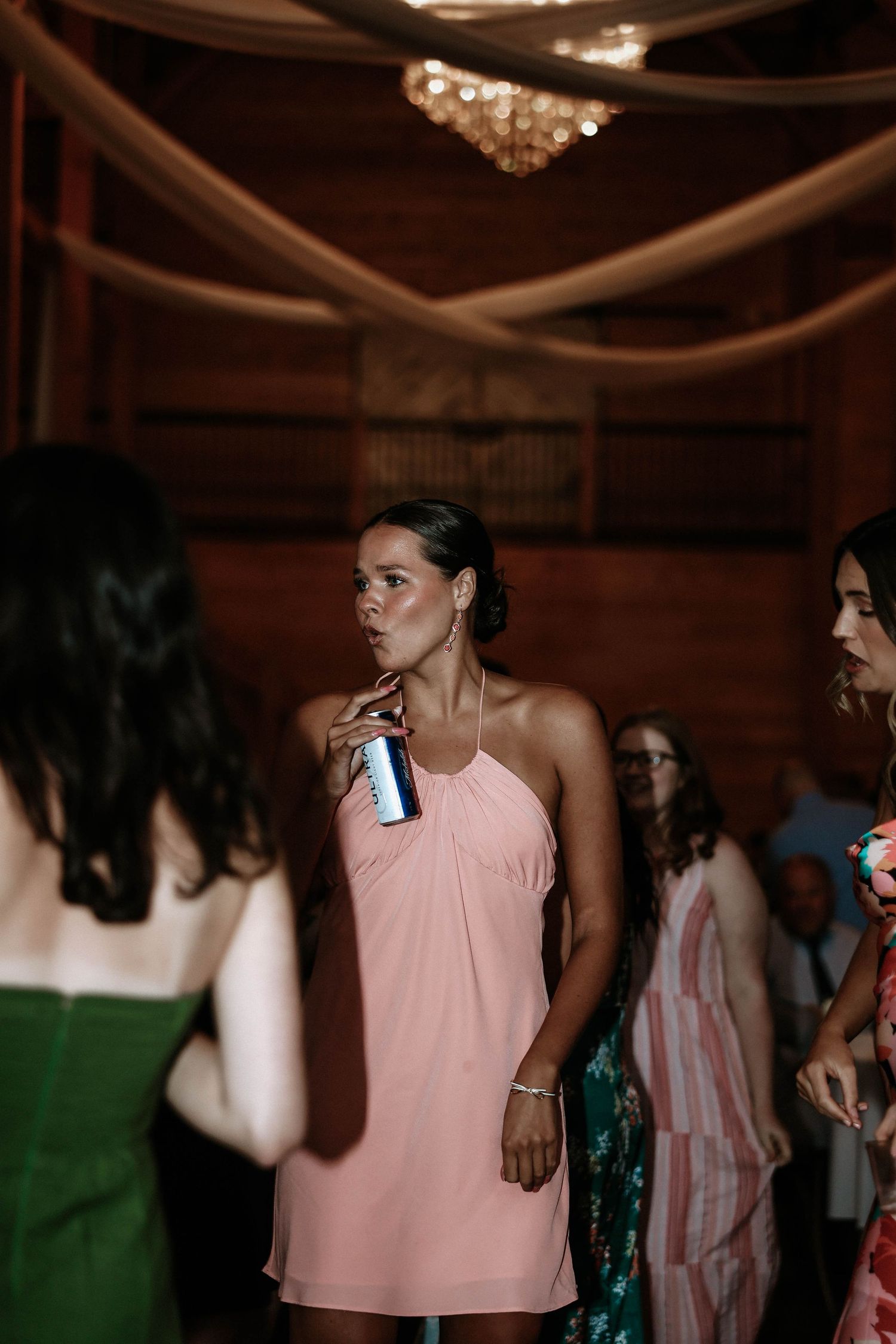 Woman in pink halter dress holding a drink can at an elegant barn wedding reception party.