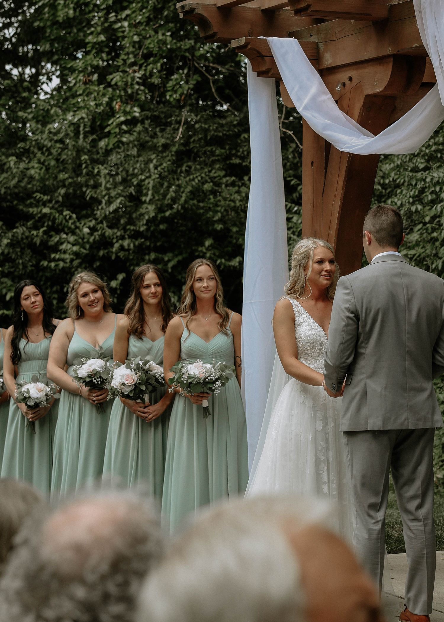 Bride and bridesmaids in sage green dresses holding bouquets at an outdoor wedding ceremony under a wooden arch.