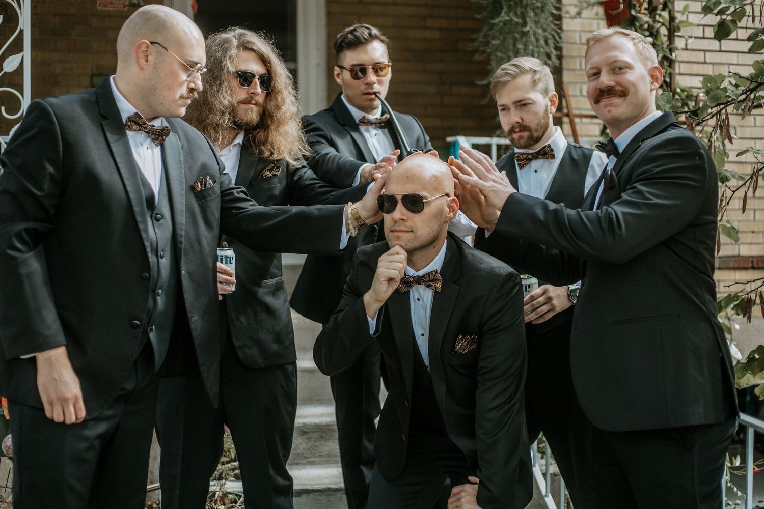 Groom and groomsmen in black tuxedos posing playfully outdoors before wedding ceremony.