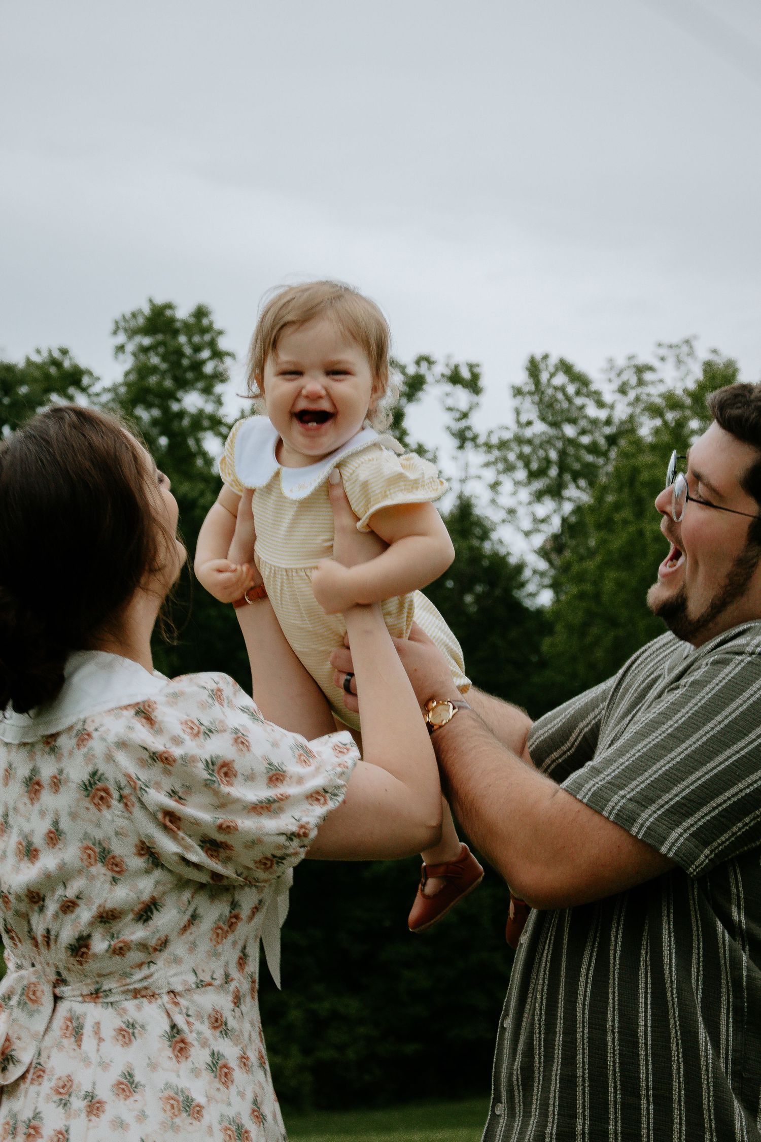 Joyful baby girl in yellow dress lifted by laughing parents outdoors in green park setting at French Park in Cincinnati, Ohio
