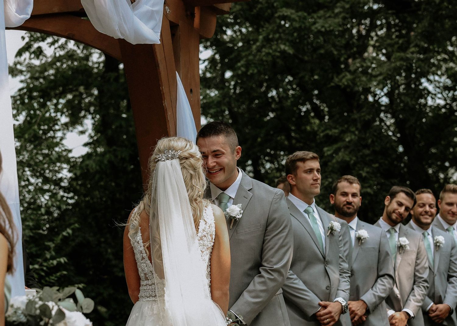Groom smiling at bride during outdoor wedding ceremony with groomsmen in gray suits at Stone Valley Meadows in Dayton, Ohio
