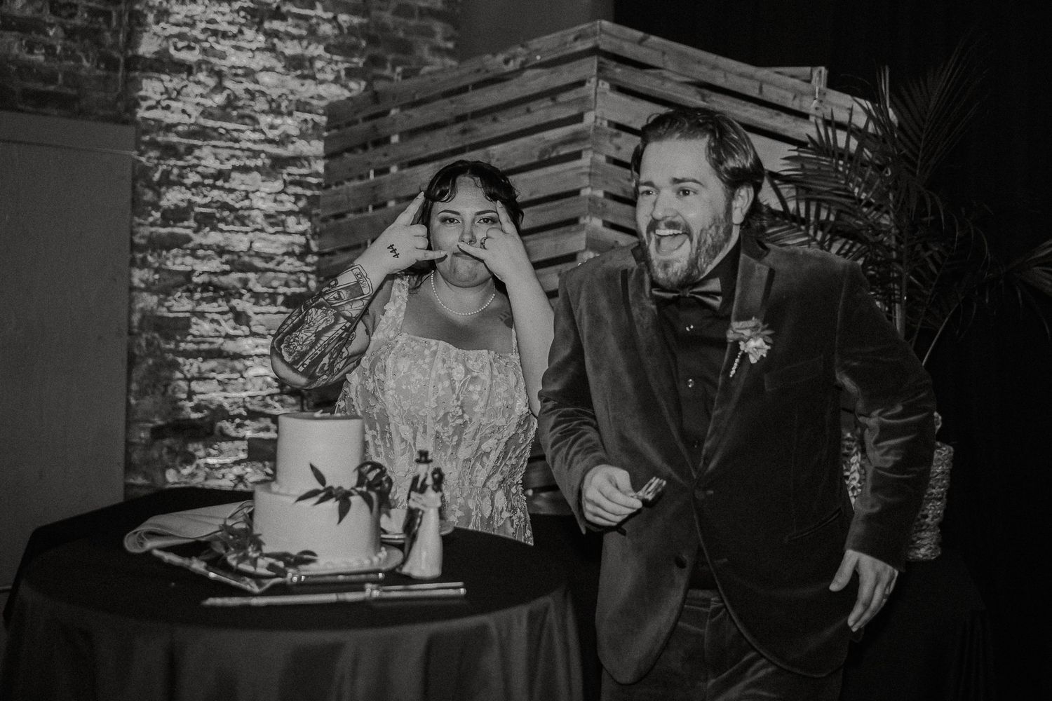Black and white photo of bride and groom laughing playfully beside their wedding cake.
