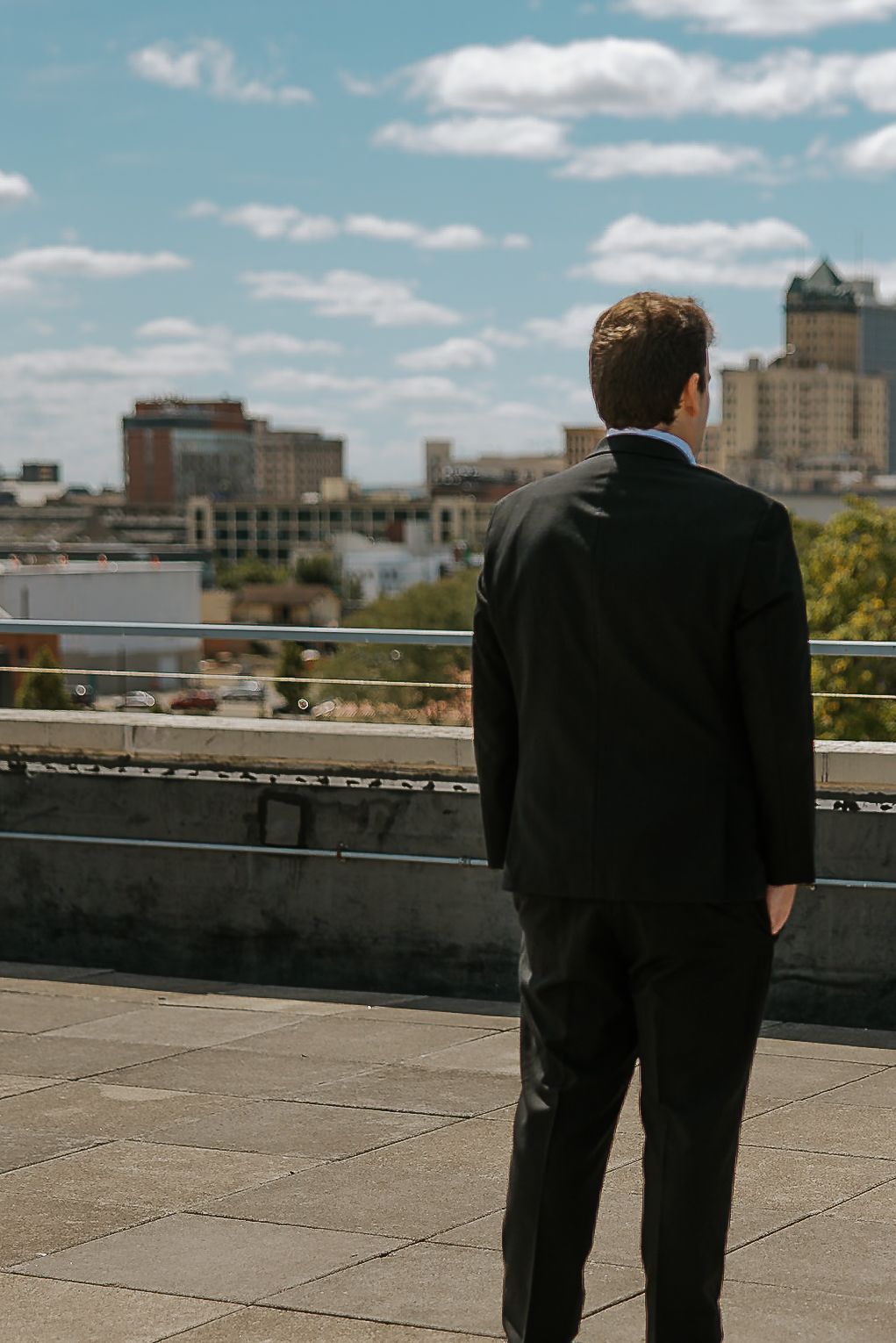 Groom in black suit standing on rooftop overlooking a sunny city skyline on wedding day.