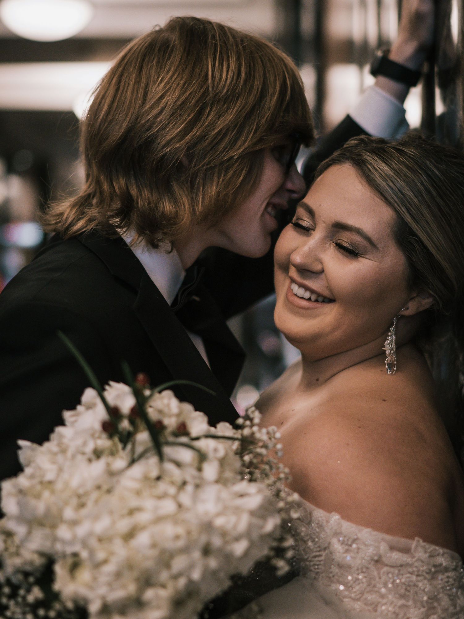 Groom kisses smiling bride on cheek as she holds white wedding bouquet in beaded off-shoulder dress.