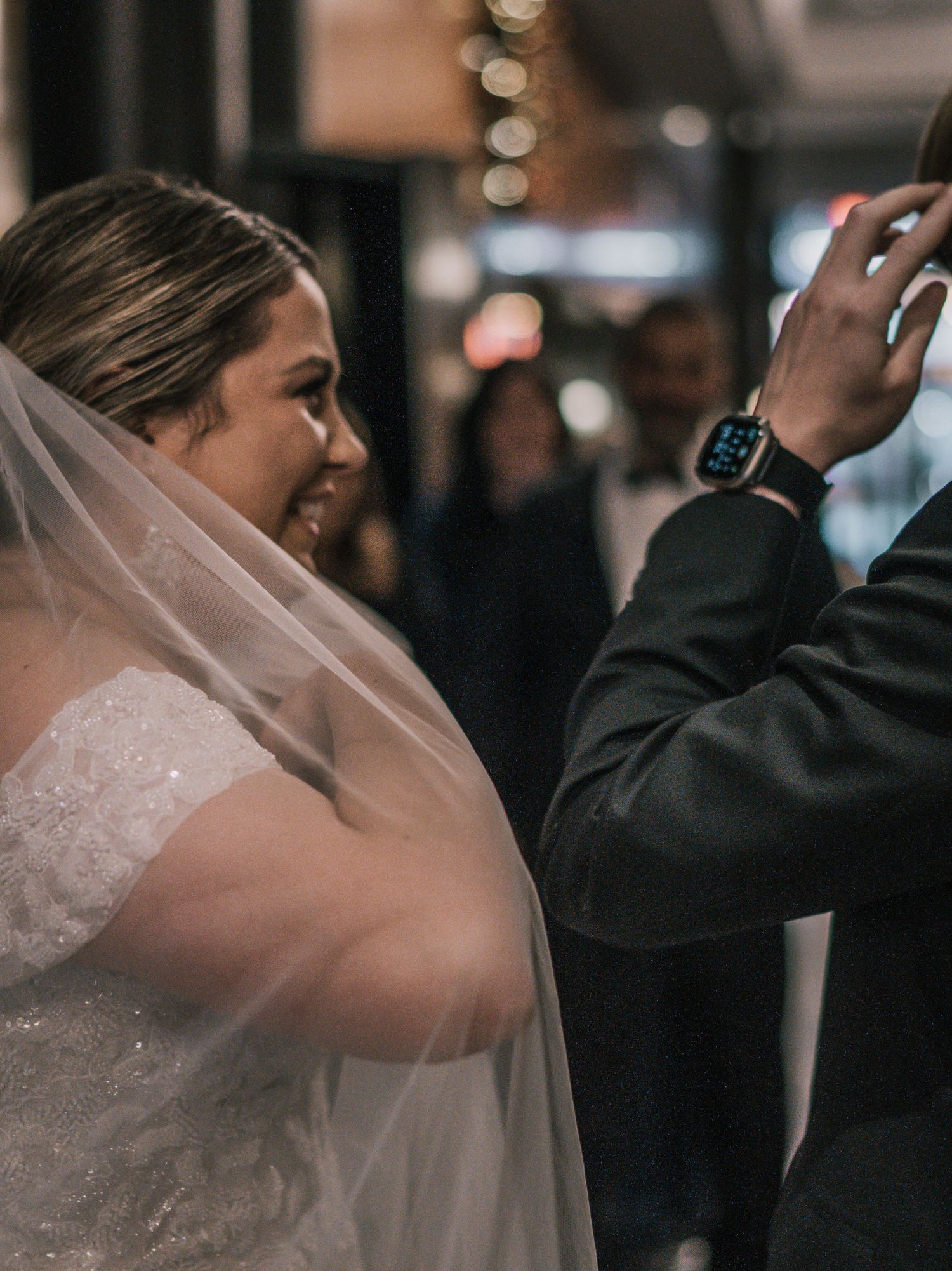 Smiling bride in white veil and gown stands beside groom wearing Apple Watch at wedding reception.