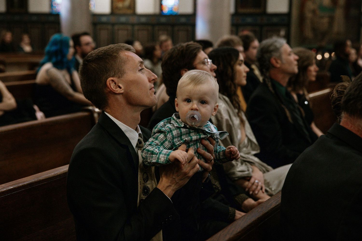 Man in black suit holds baby in plaid outfit while seated in church pew during wedding ceremony in Cincinnati, Ohio