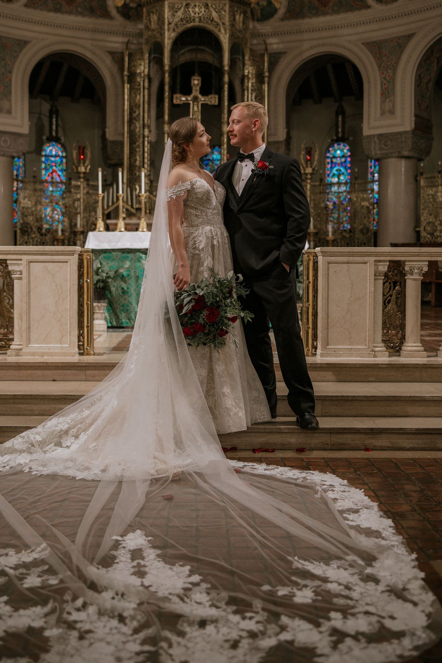Bride and groom pose at church altar showcasing stunning lace wedding gown with cathedral-length veil in Cincinnati, Ohio