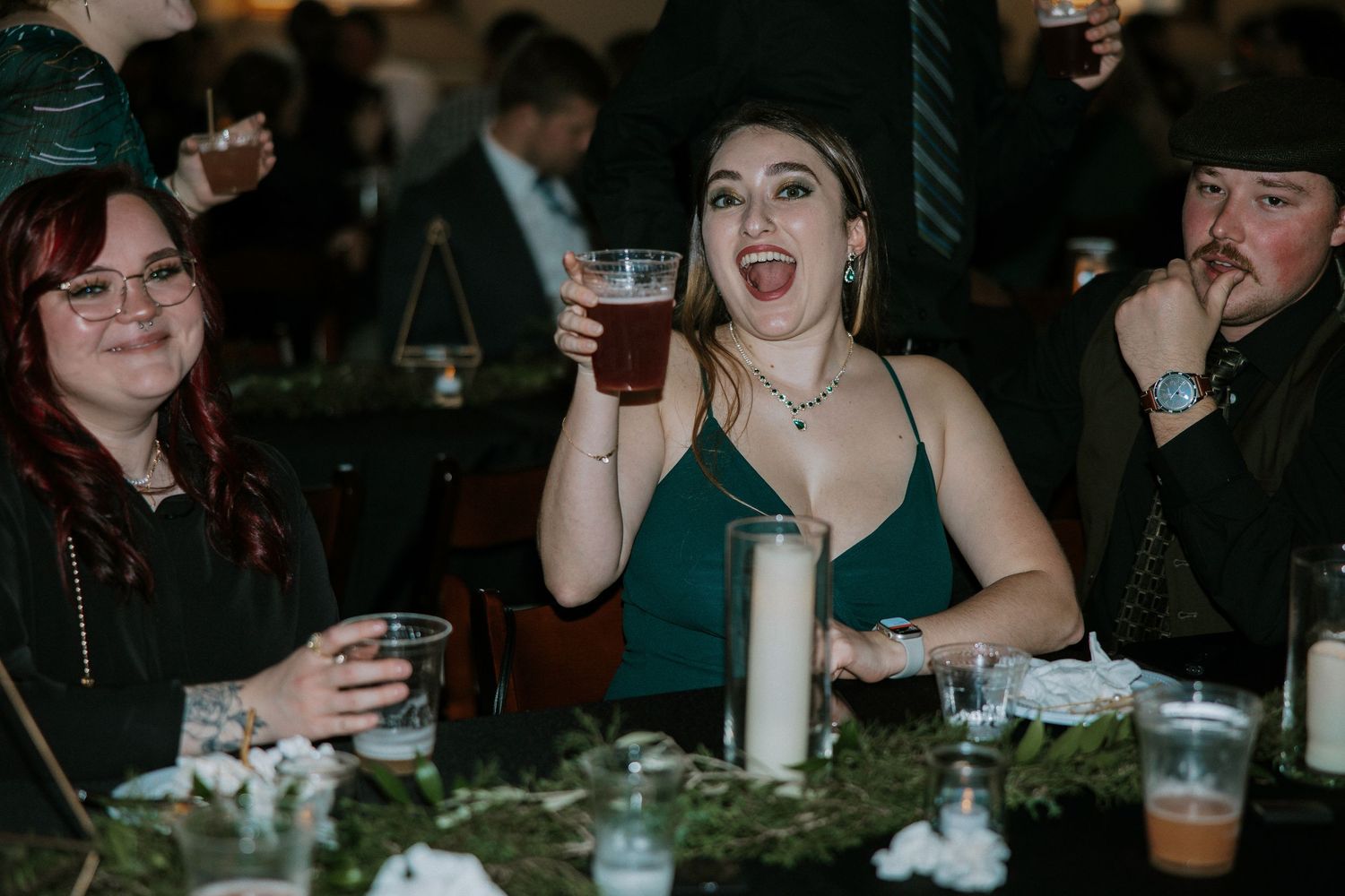 Wedding guests laughing and raising drinks at a reception dinner table with candles and greenery at Urban Artifact in Cincinnati, Ohio