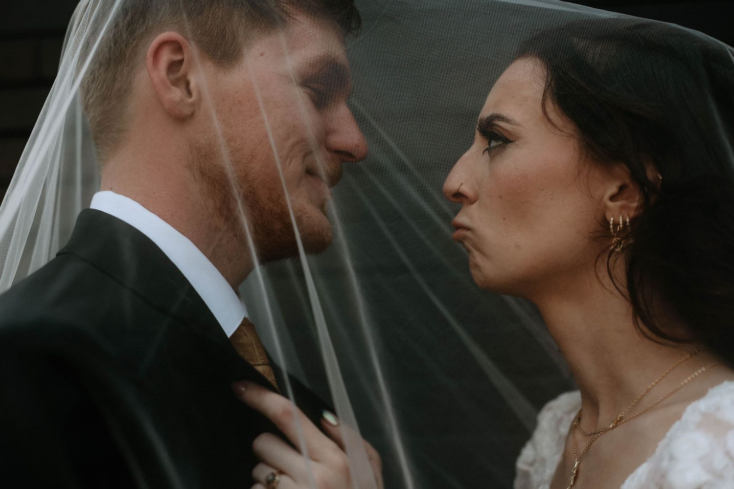 Bride and groom share a romantic moment under a wedding veil, faces close together in an intimate pose.