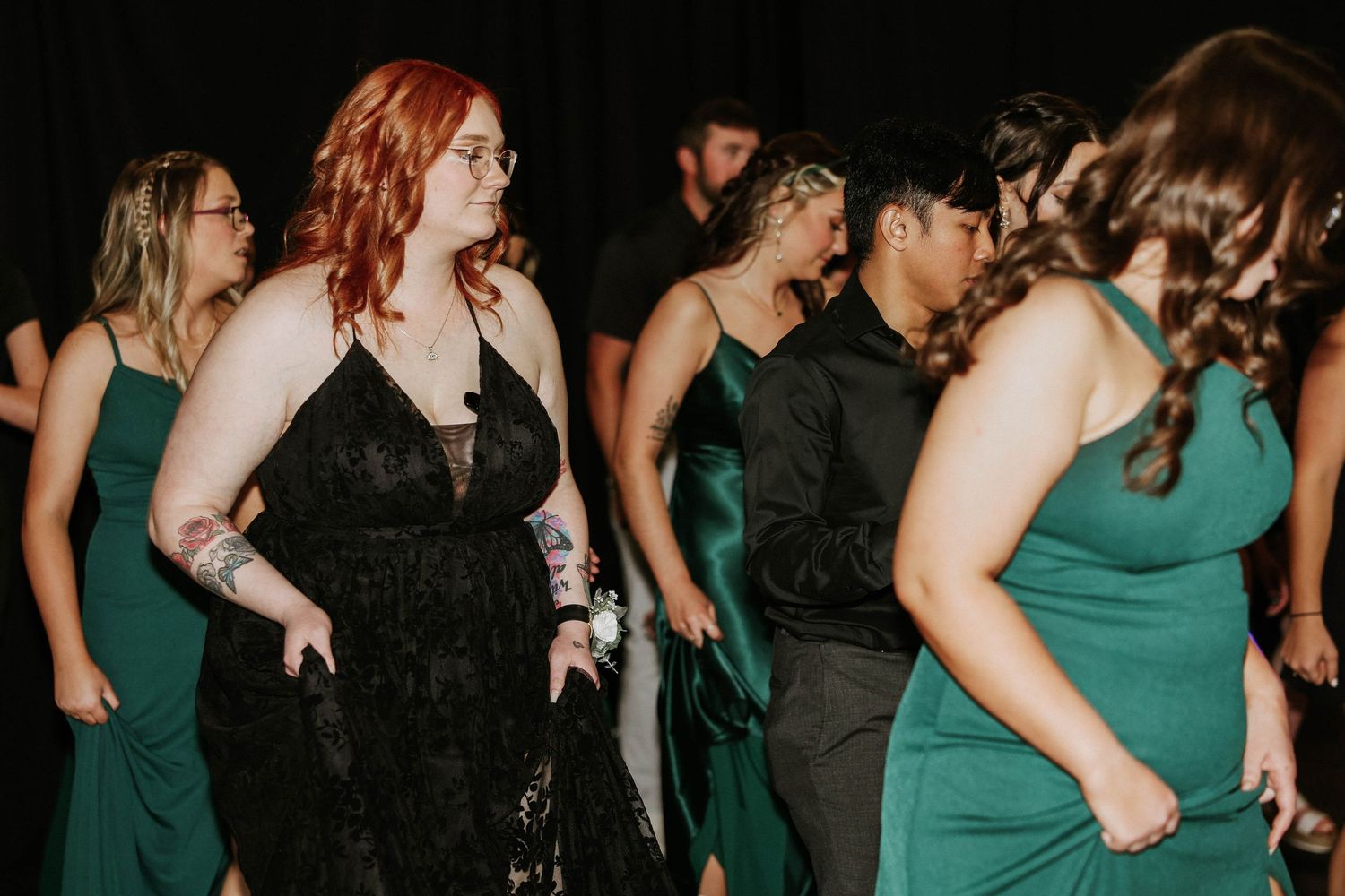 Group of women in formal gowns dancing at prom or gala event, wearing green and black dresses.
