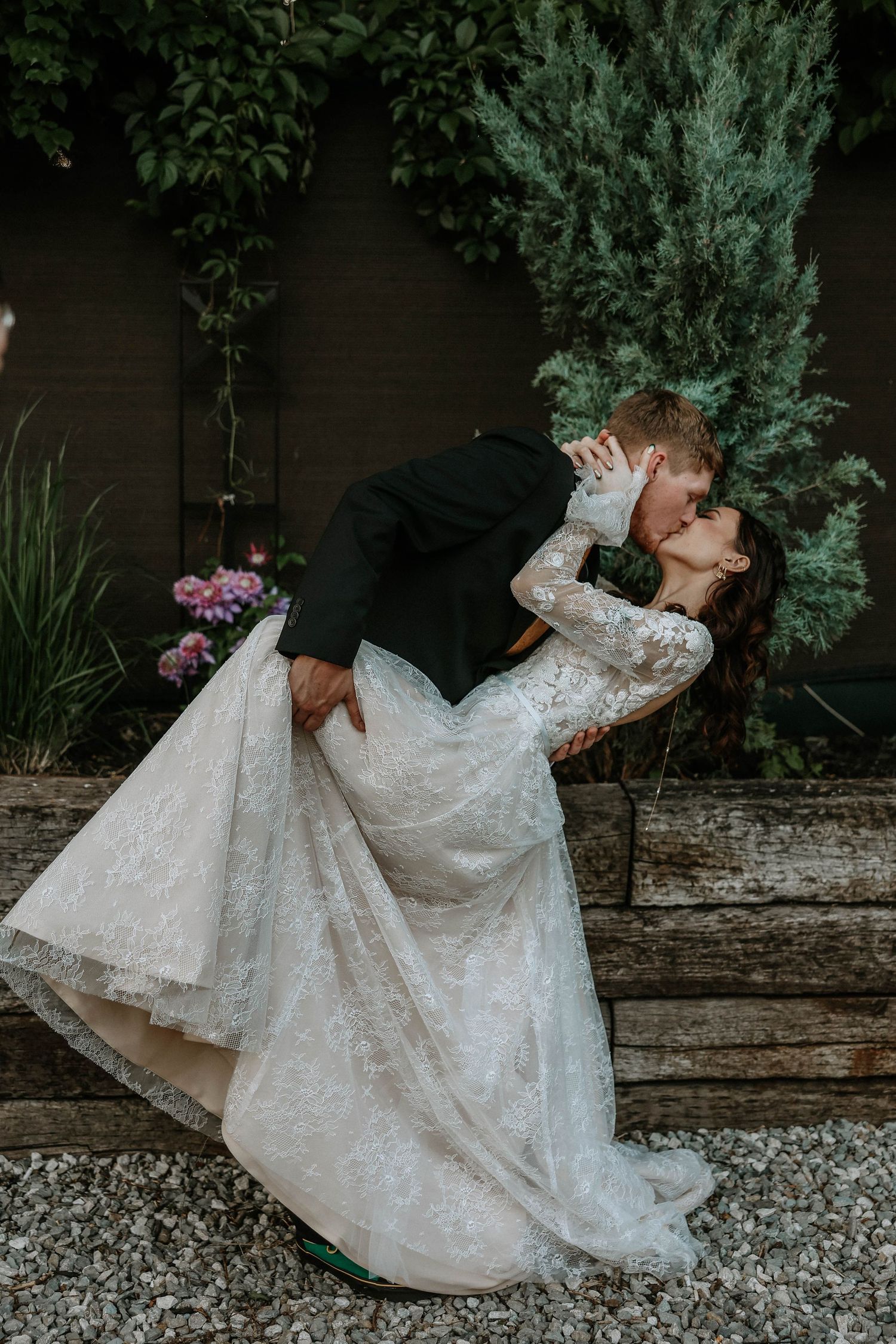 Groom dips bride in a romantic kiss outdoors, bride wearing a lace long-sleeve wedding gown surrounded by lush greenery.