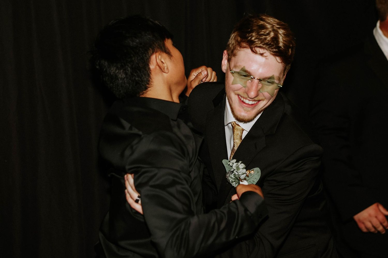 Two young men in formal black suits share a friendly embrace at a prom or formal event.