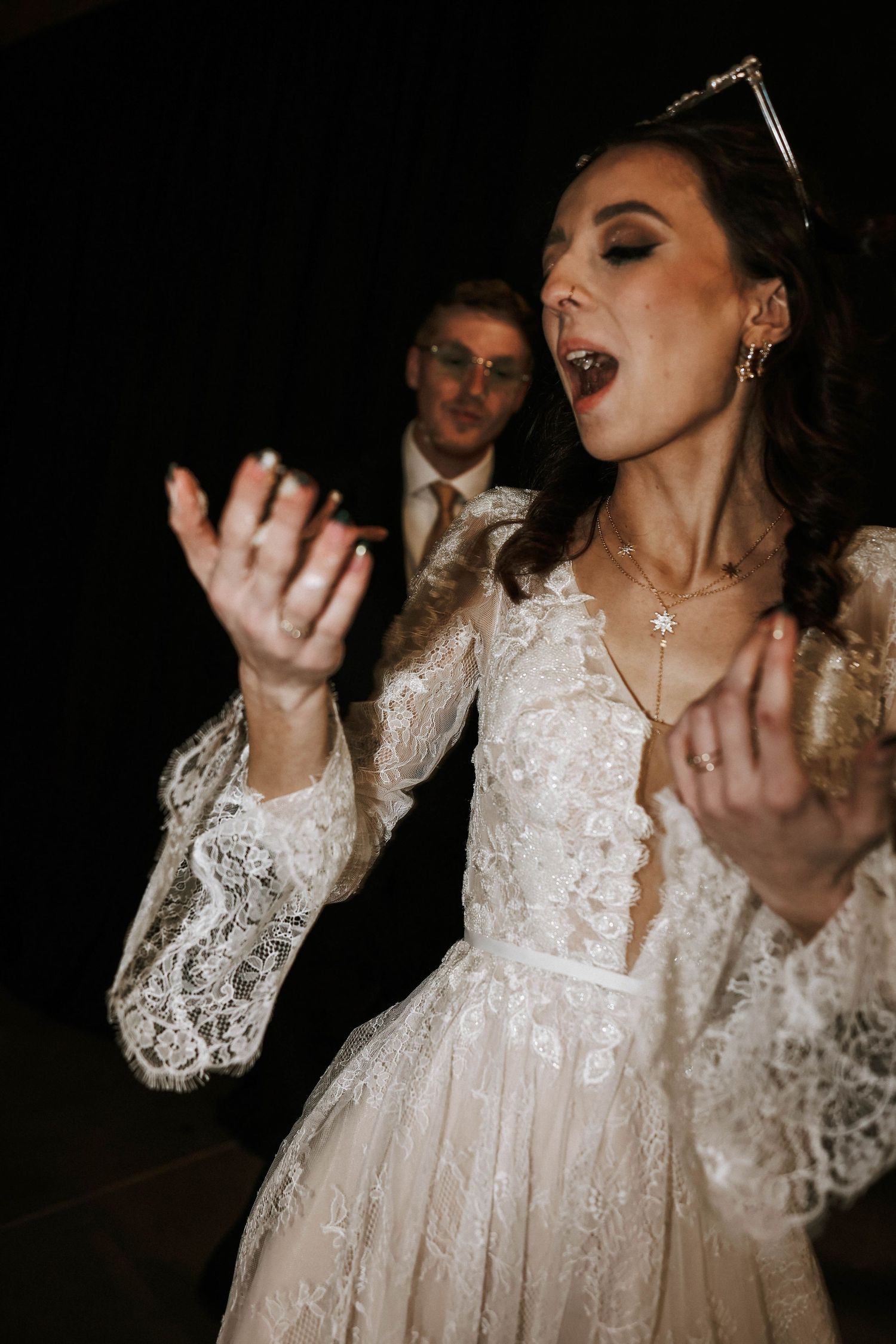 Bride in lace wedding dress dancing expressively at wedding reception with groom in background.