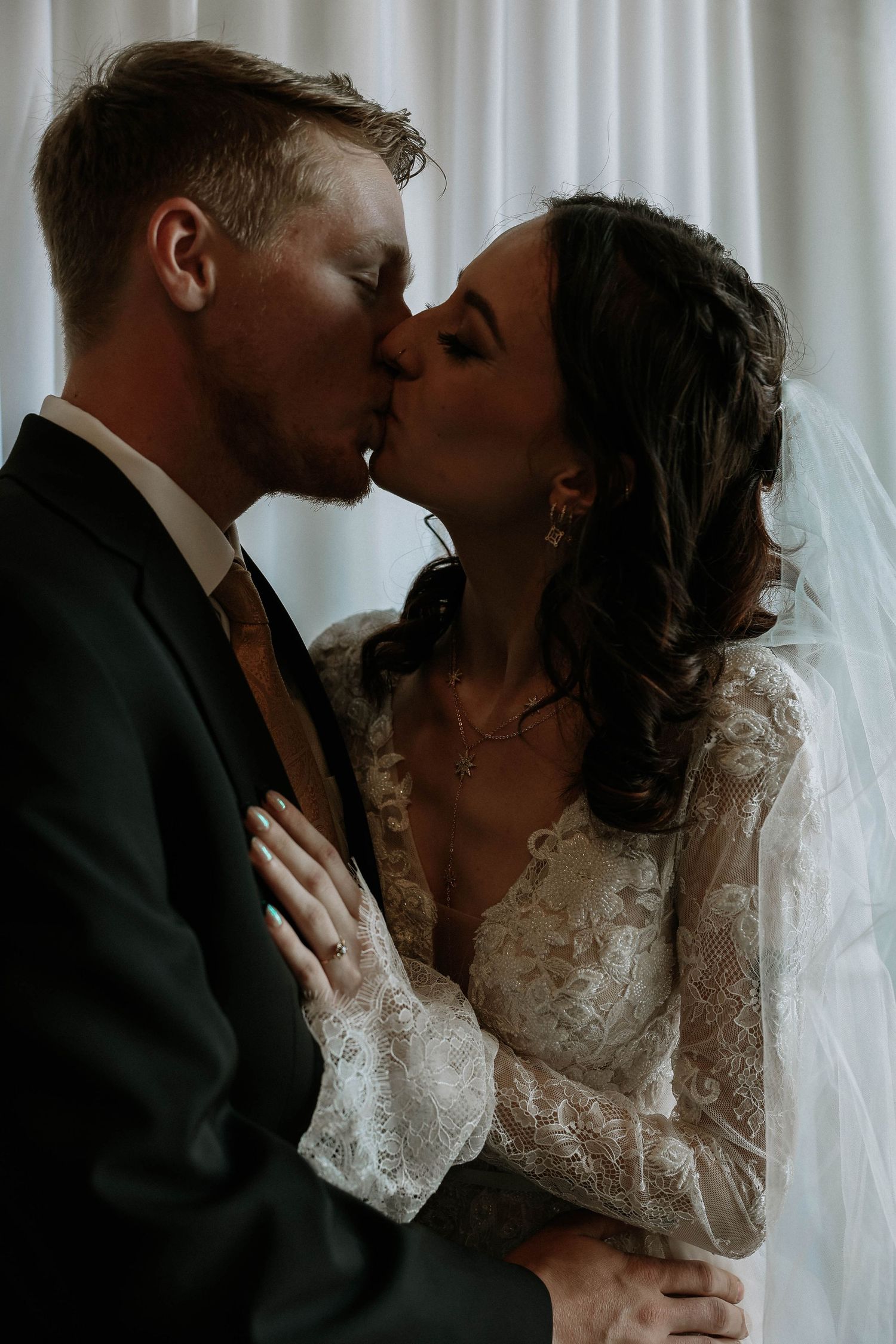 A bride and groom share a romantic kiss in soft light, she wearing a lace long-sleeve gown and veil.