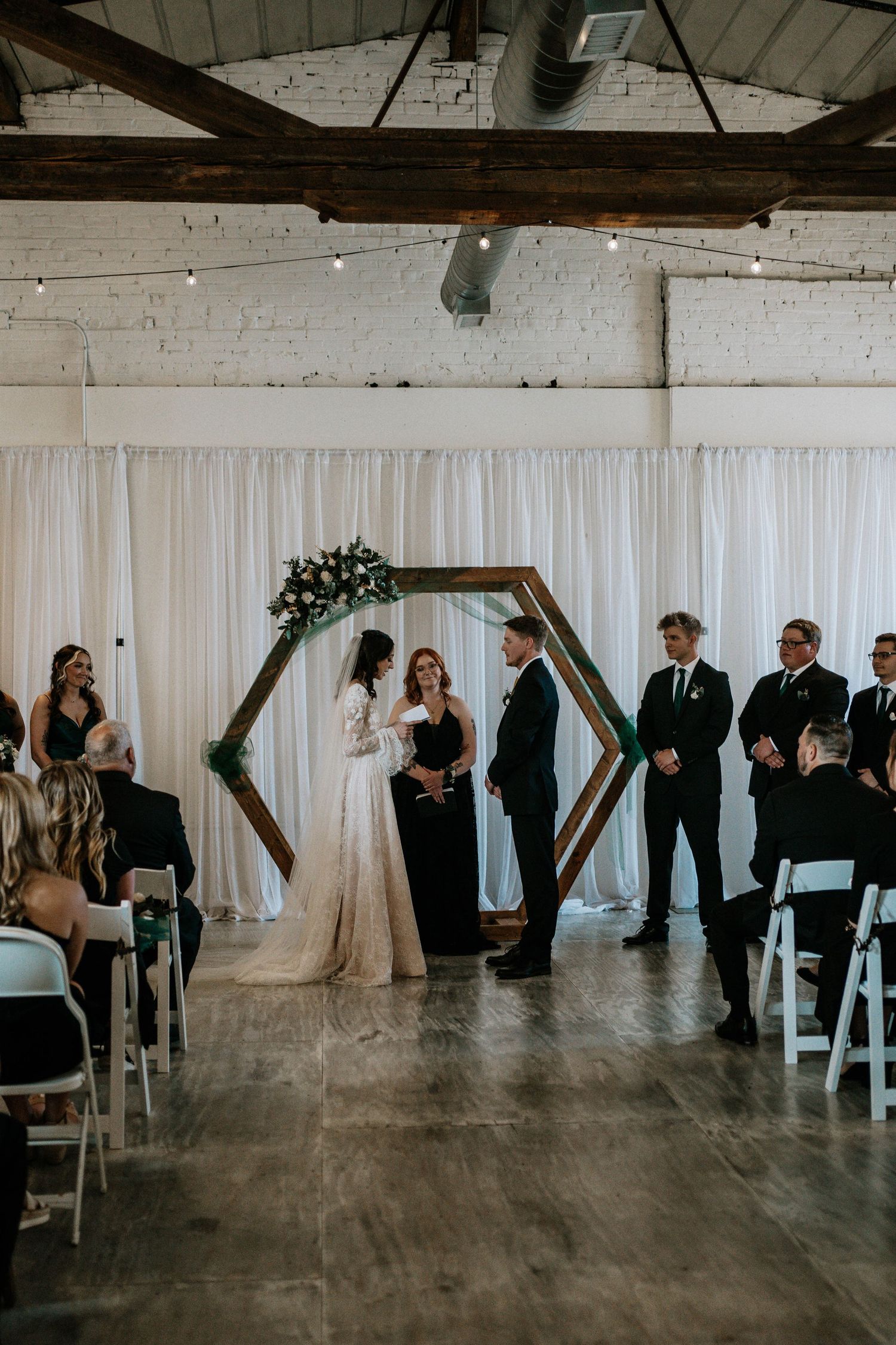 Couple exchanges vows under a hexagonal wooden arch with floral accents in an elegant indoor wedding ceremony.