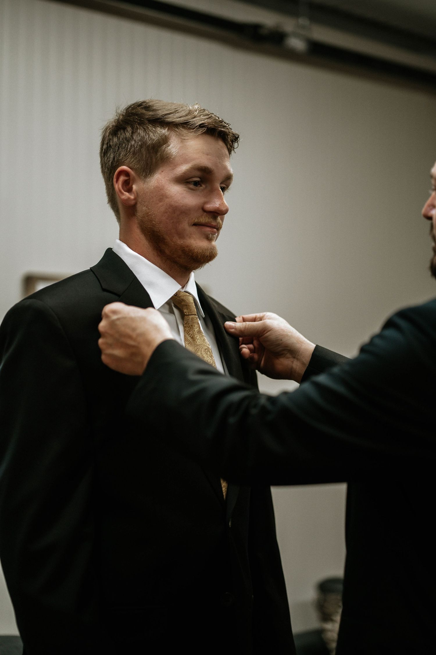 A groom in a black suit and gold tie gets his lapel adjusted by a groomsman before the wedding ceremony.