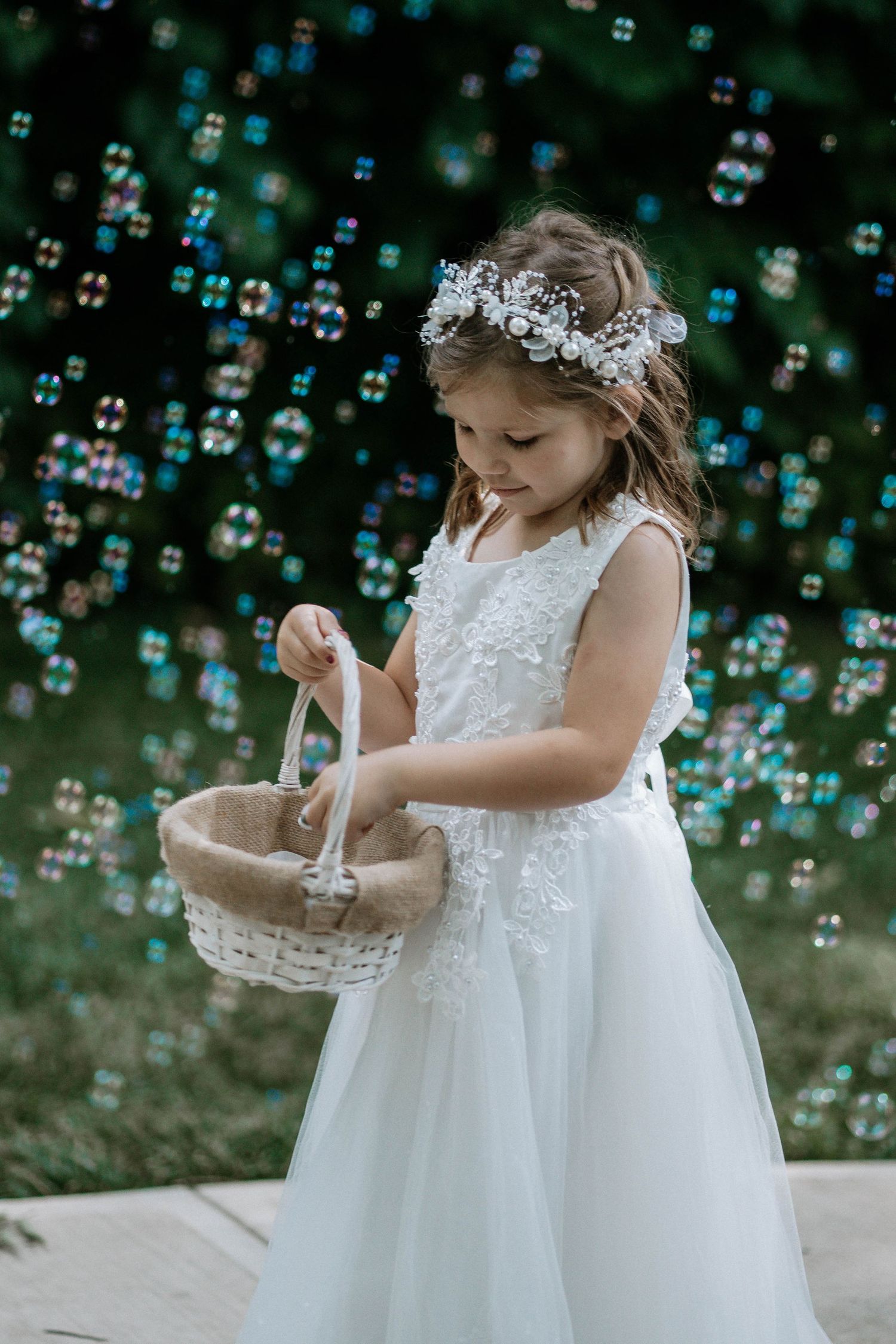 Flower girl in white lace dress with floral crown holds a basket surrounded by colorful soap bubbles.