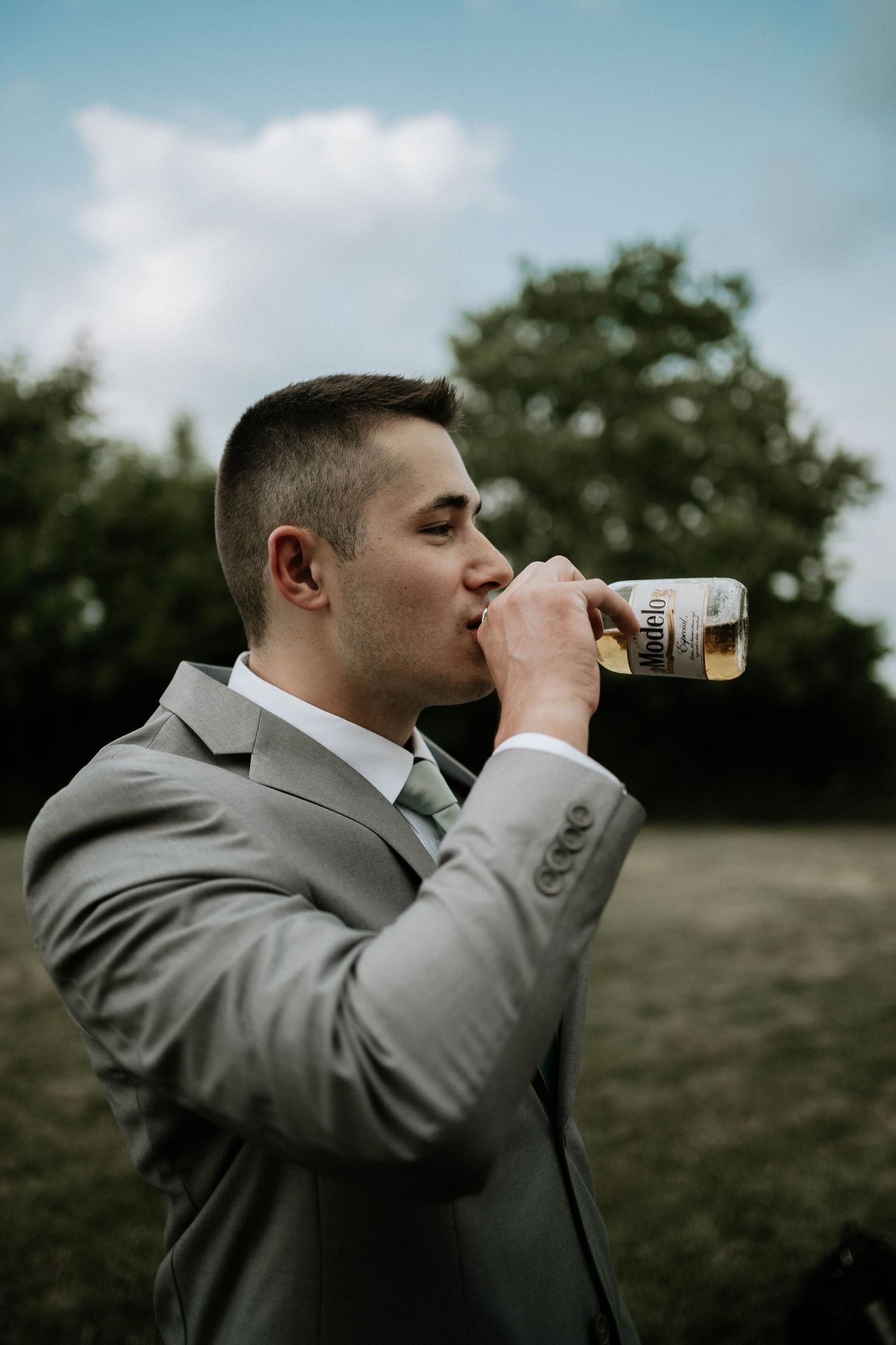 Groom in gray suit drinking from a beer bottle outdoors with green trees in the background on wedding day.