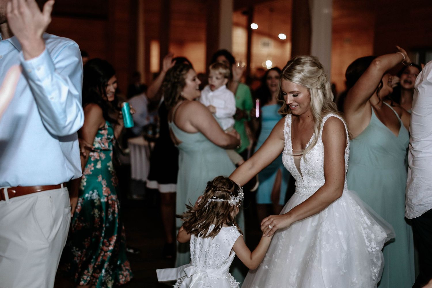 Bride in white gown dances joyfully with flower girl on wedding reception dance floor.