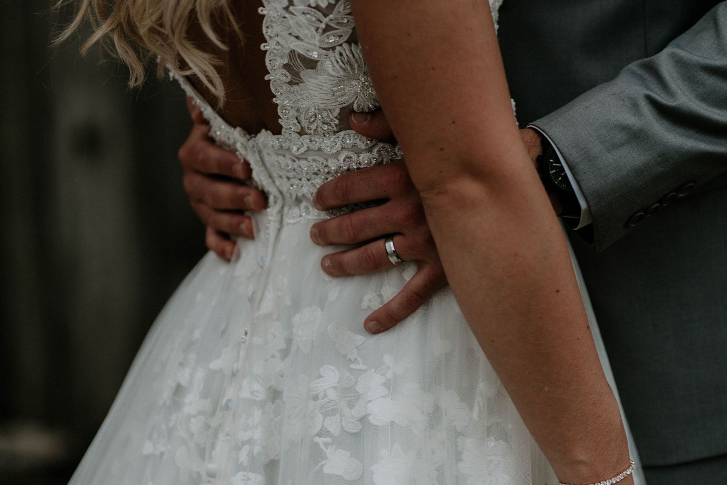 Close-up of a bride in a lace wedding dress with groom's hand on her waist, wearing a wedding ring.